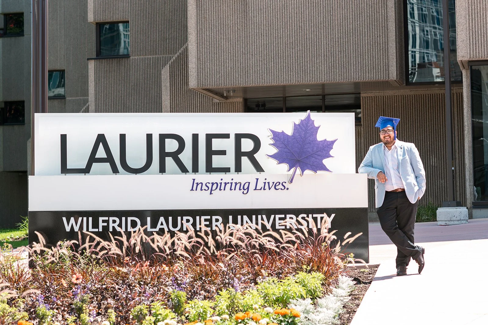 Lyndsi Seth photography- A male graduate in a suit jacket and blue cap leaning against the Wilfrid Laurier University sign on campus
