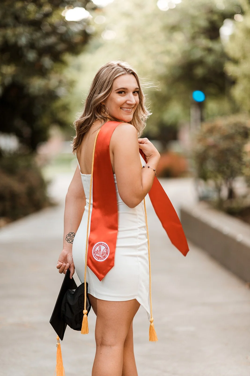 Lyndsi Seth Photography-A smiling graduate looking back over her shoulder outdoors while wearing a white dress, a red graduation stole, and holding her black cap