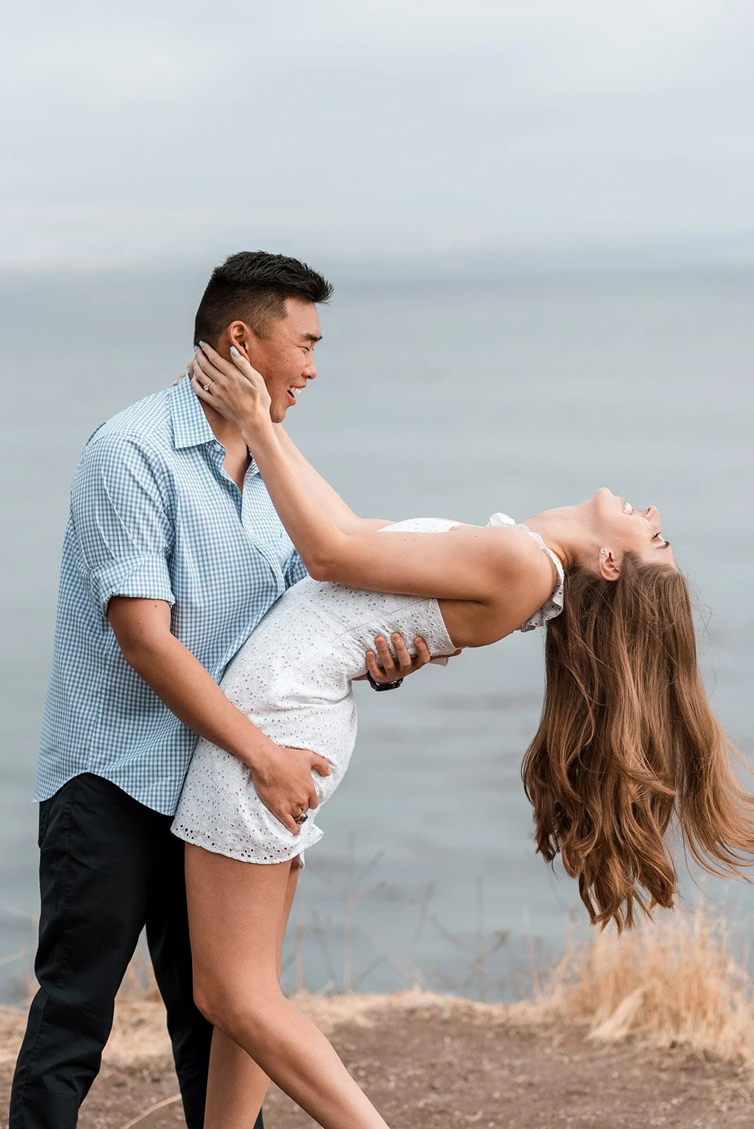 A man holding a woman in a dip pose near a body of water, both smiling and enjoying the moment.