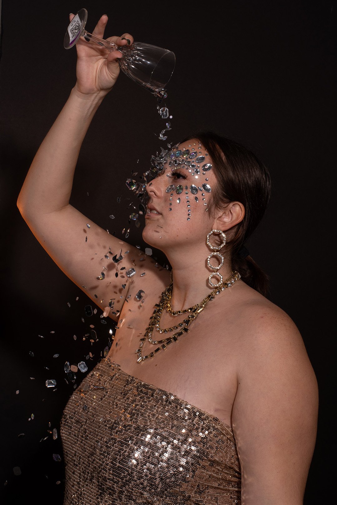 Lyndsi Seth photography- A dramatic editorial portrait of a woman with her arm raised, adorned with chunky silver and gold face jewels around her eye and wearing layered gold chain necklaces
