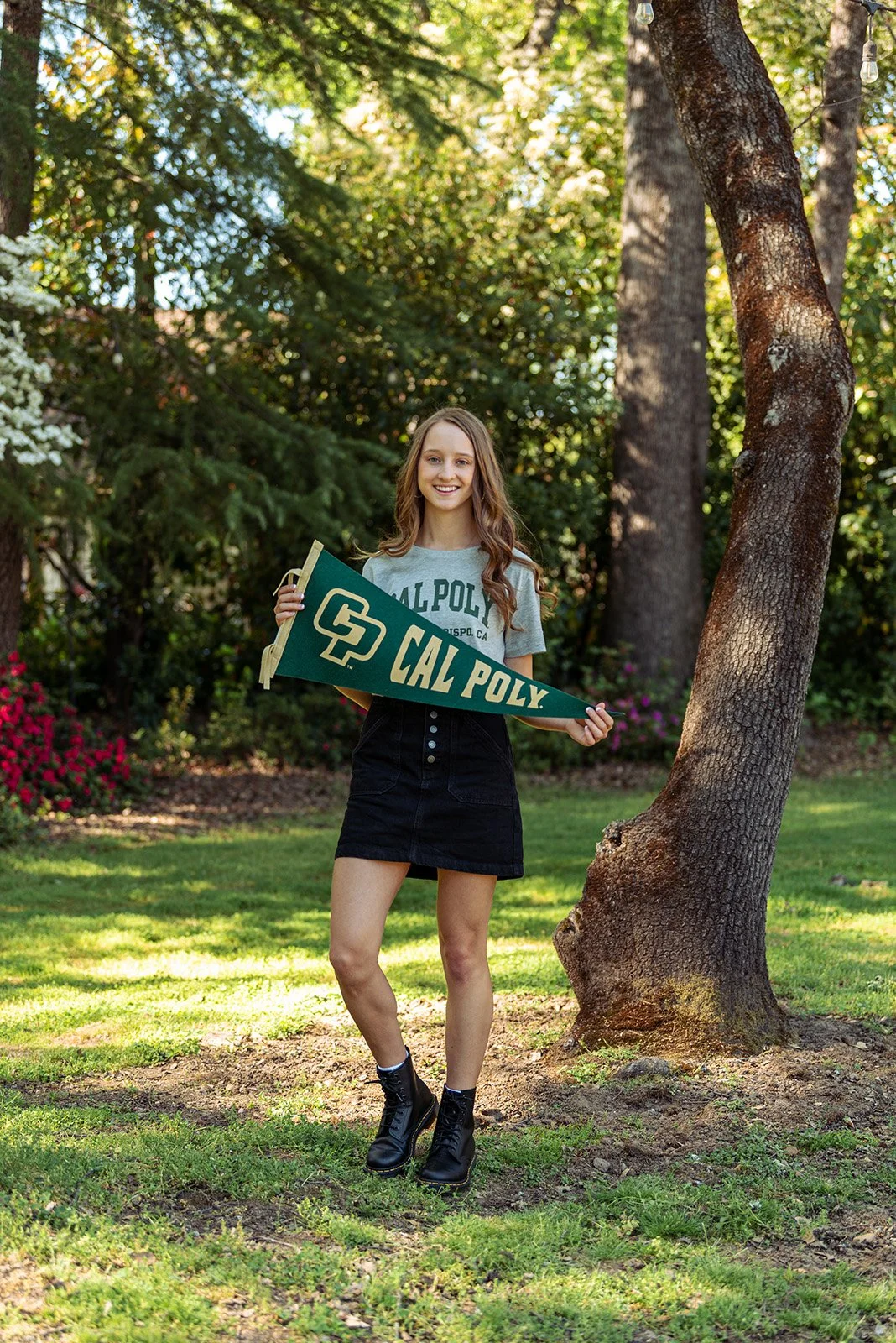 A young woman standing outdoors on grass, holding a green San Diego Cal Poly pennant, smiling at the camera. She is wearing a Cal Poly T-shirt, a black skirt, and black combat boots. There are trees and bushes in the background.