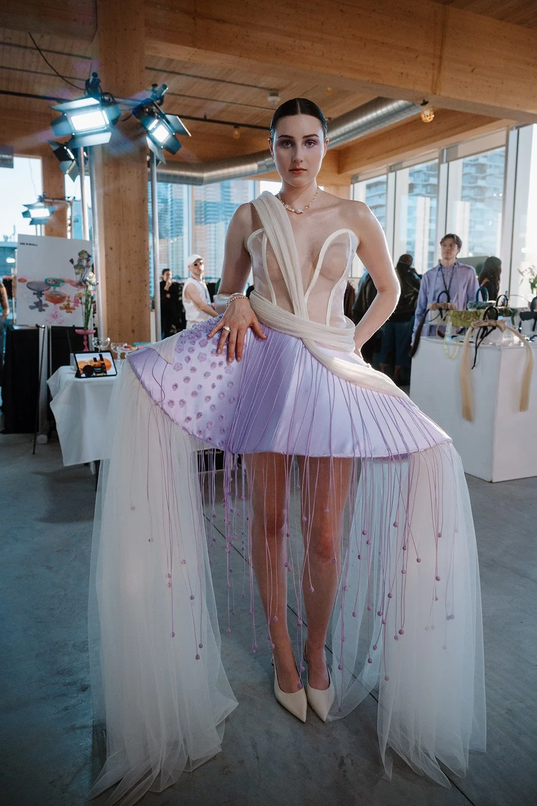 Lyndsi Seth photography- A fashion model wearing an avant-garde white and lavender pleated dress with sheer trailing fabric, posing indoors during a runway event