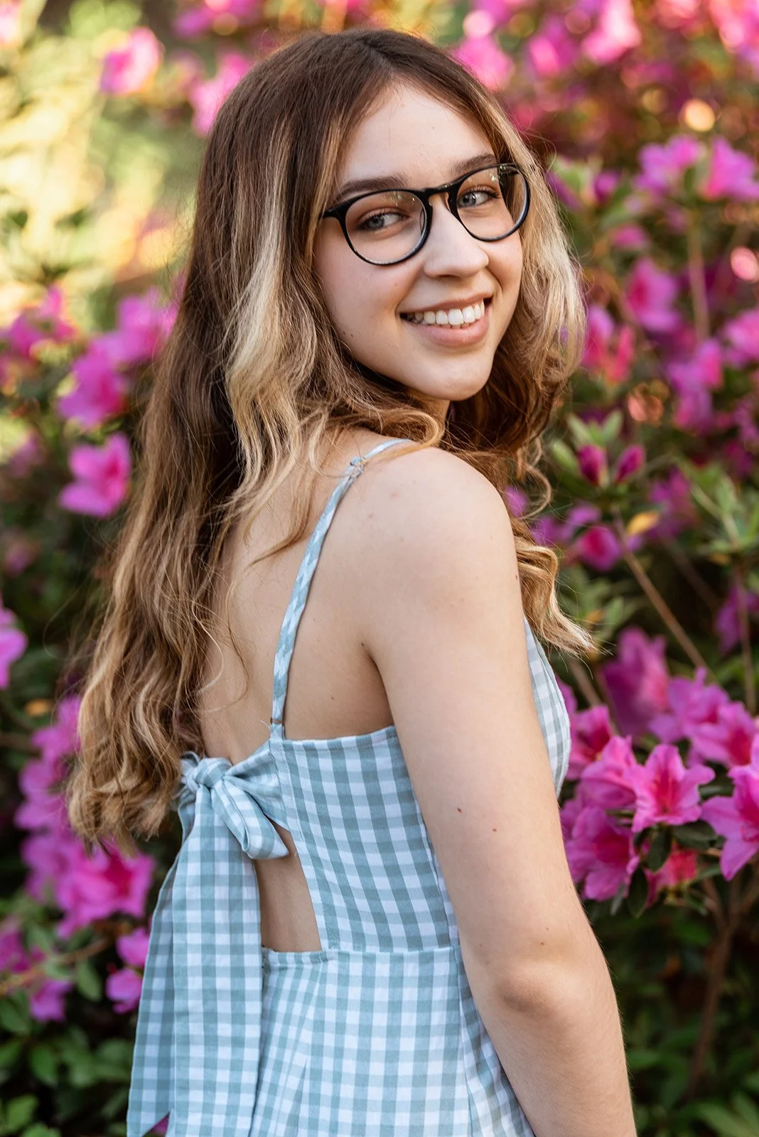 A young woman with long, curly brown hair and glasses, smiling and looking over her shoulder, standing in front of pink flowers.