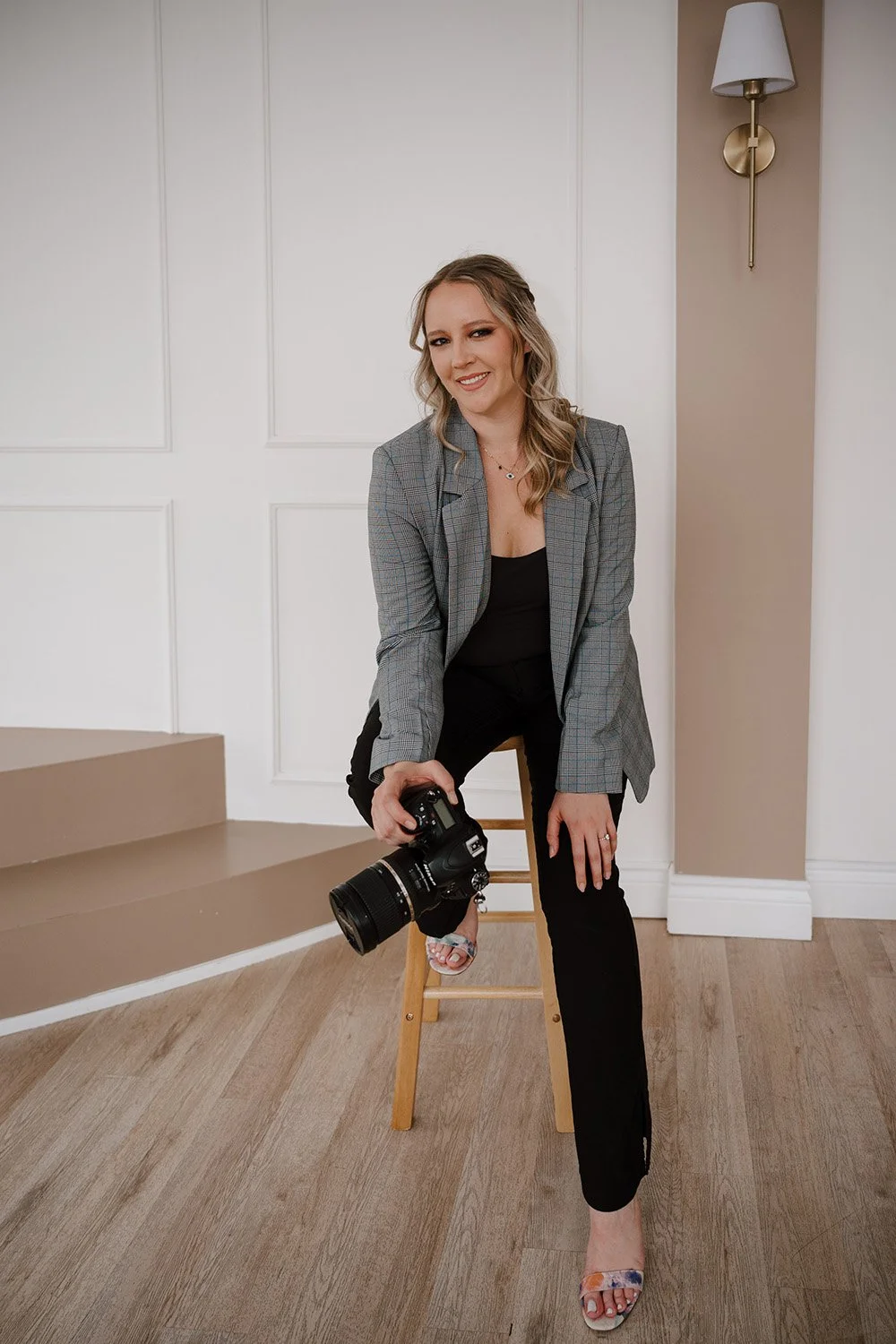 A woman with wavy blonde hair, wearing a gray plaid blazer, black top, and black pants, is sitting on a wooden stool indoors. She is holding a professional camera with her right hand and smiling at the camera. The background includes white paneled walls, a beige wall with a modern wall sconce, and wooden flooring.
