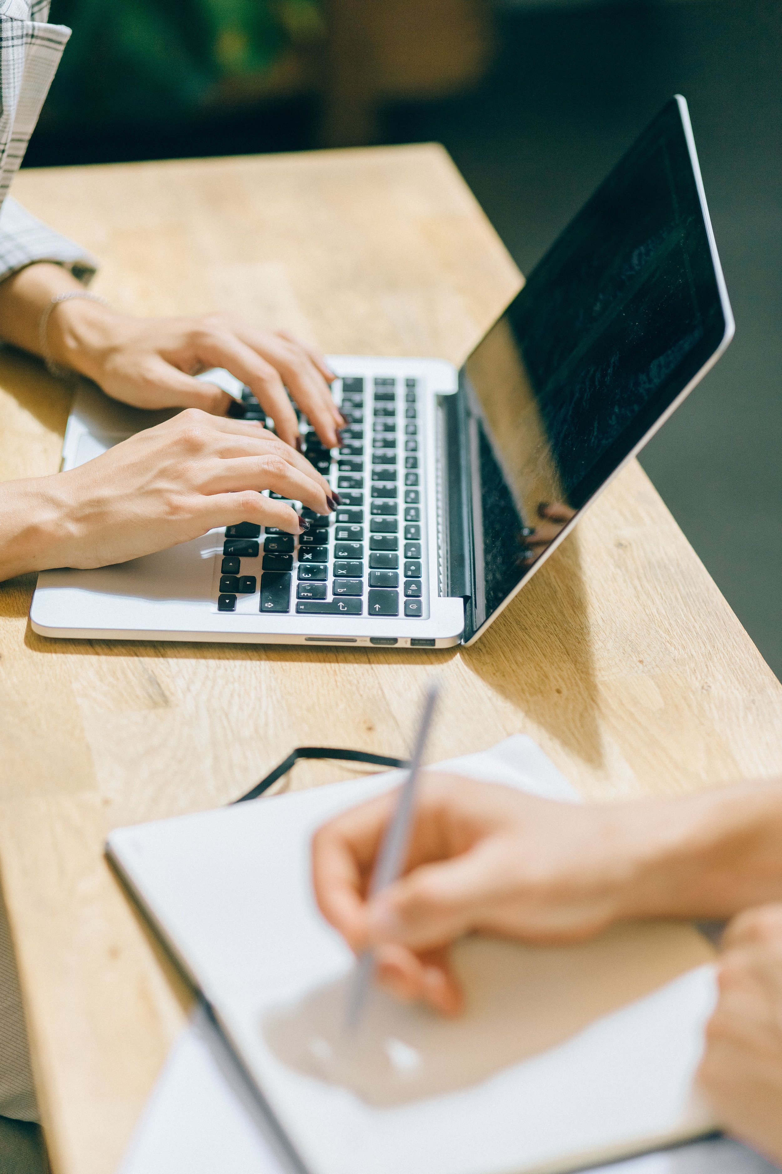 Hands typing on a laptop while a colleague writes notes at a desk.