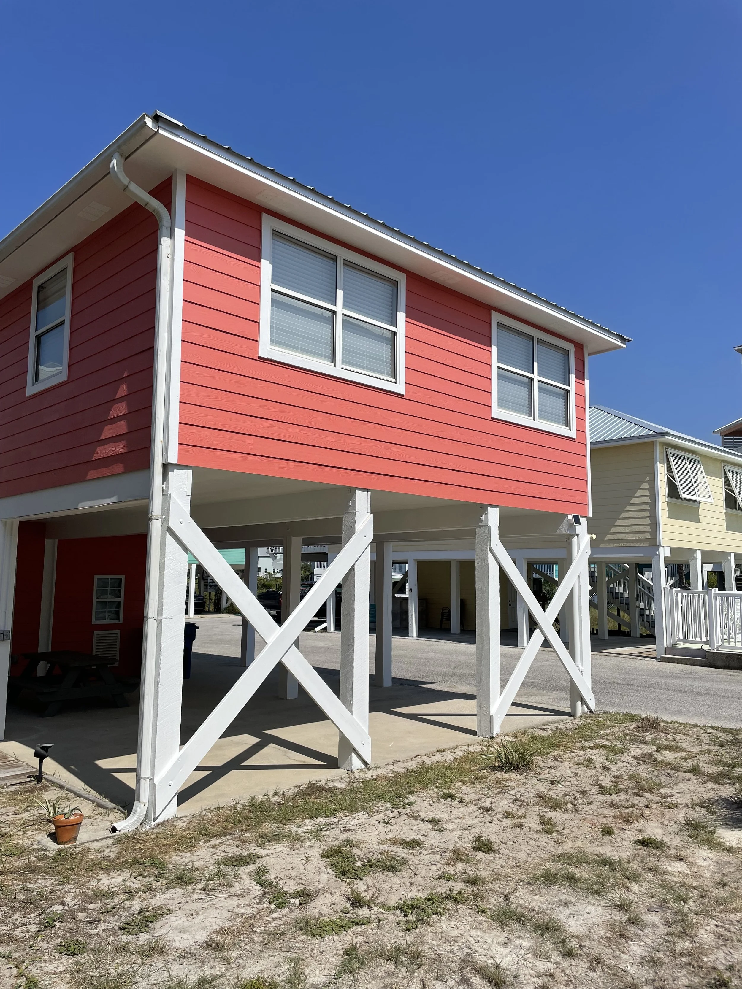 Two-story colorful house with pink siding, white trim, and metal roof, elevated on stilts with open space underneath, located in a sunny area with a sandy yard and sparse grass.