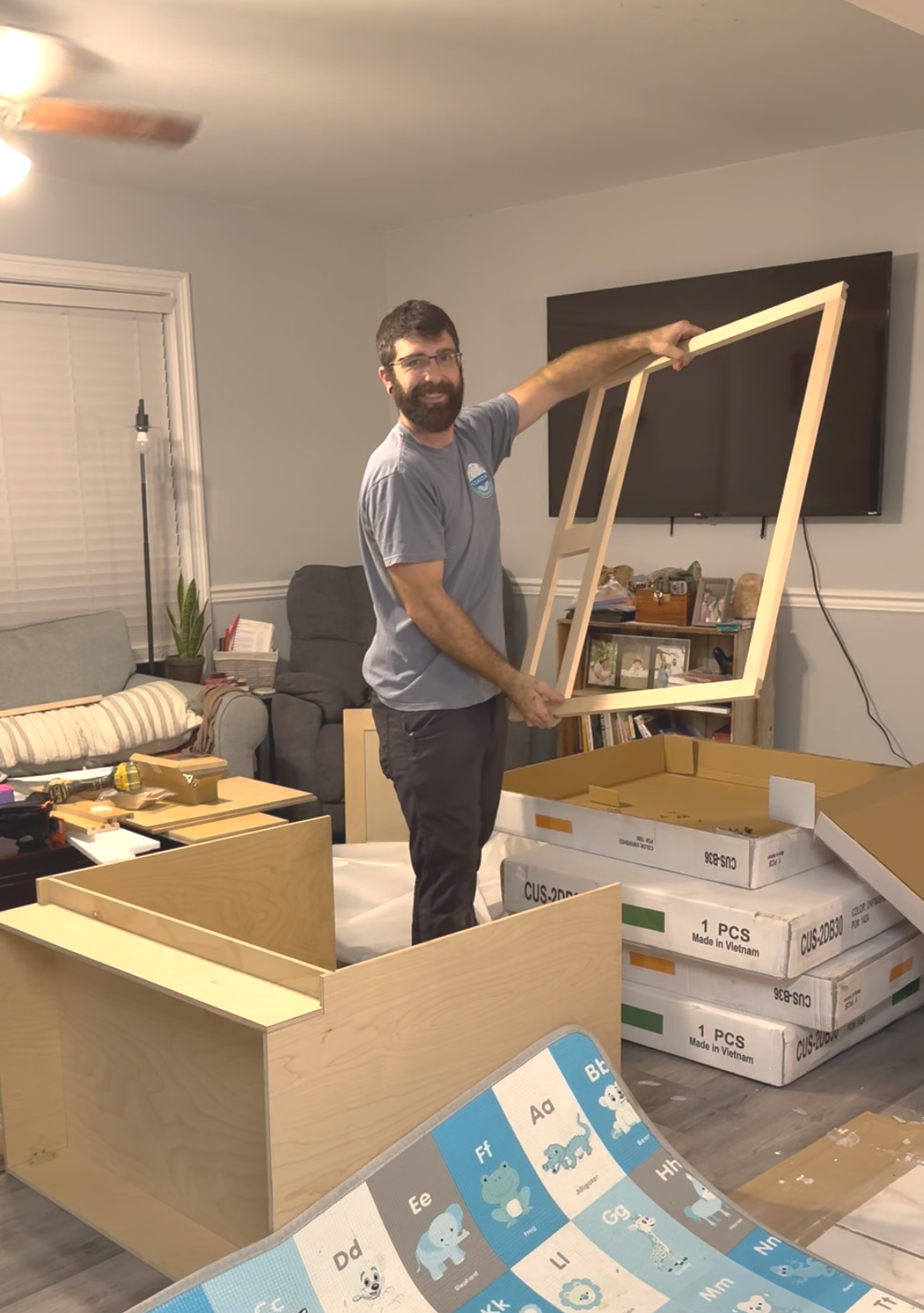A man assembling a wooden frame in a living room, with furniture, boxes, and a television on the wall behind him.