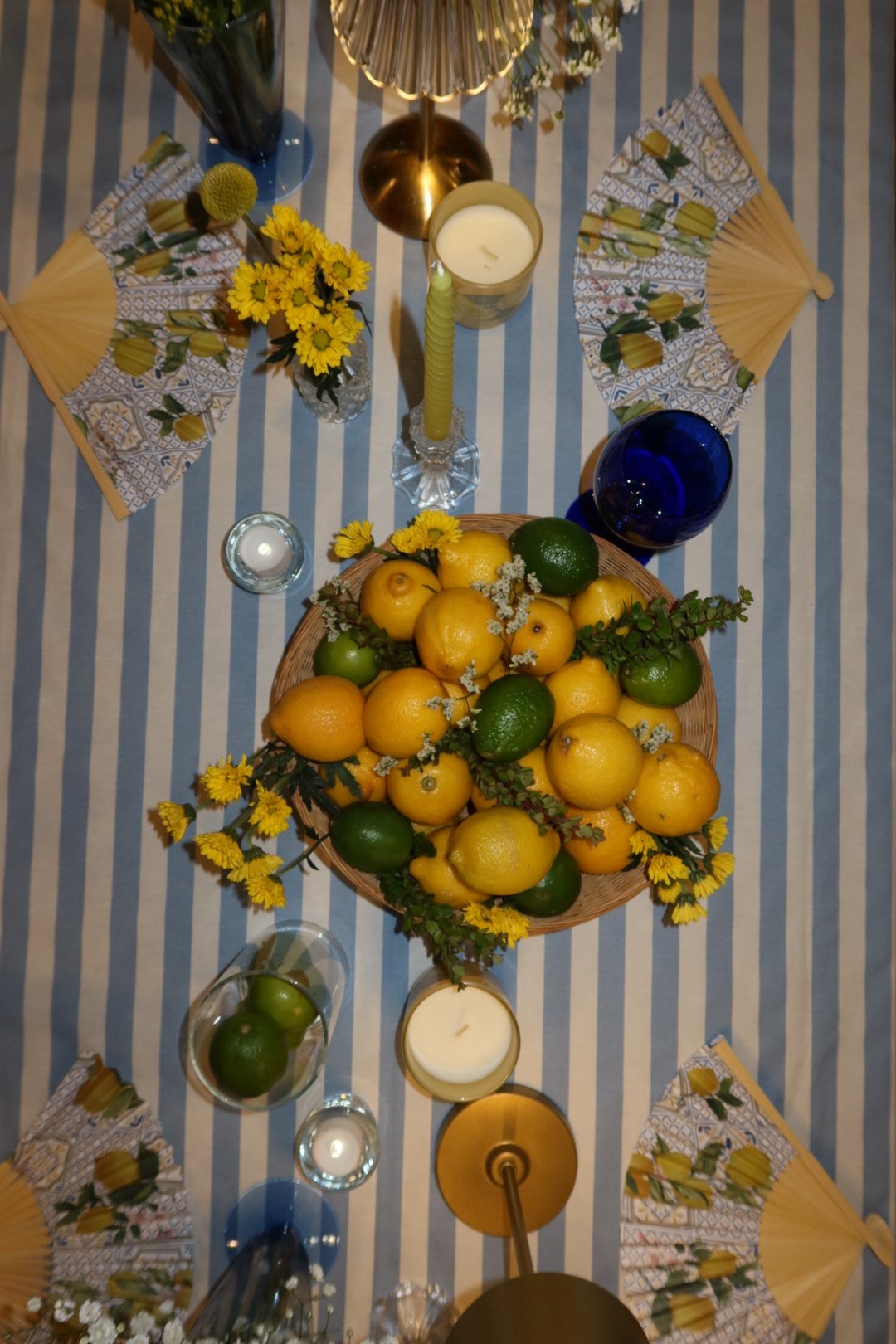 A table decorated with a blue and white striped tablecloth, featuring a large basket of lemons and limes, several candles, glasses, and floral decorations, with fans and napkins around the table.