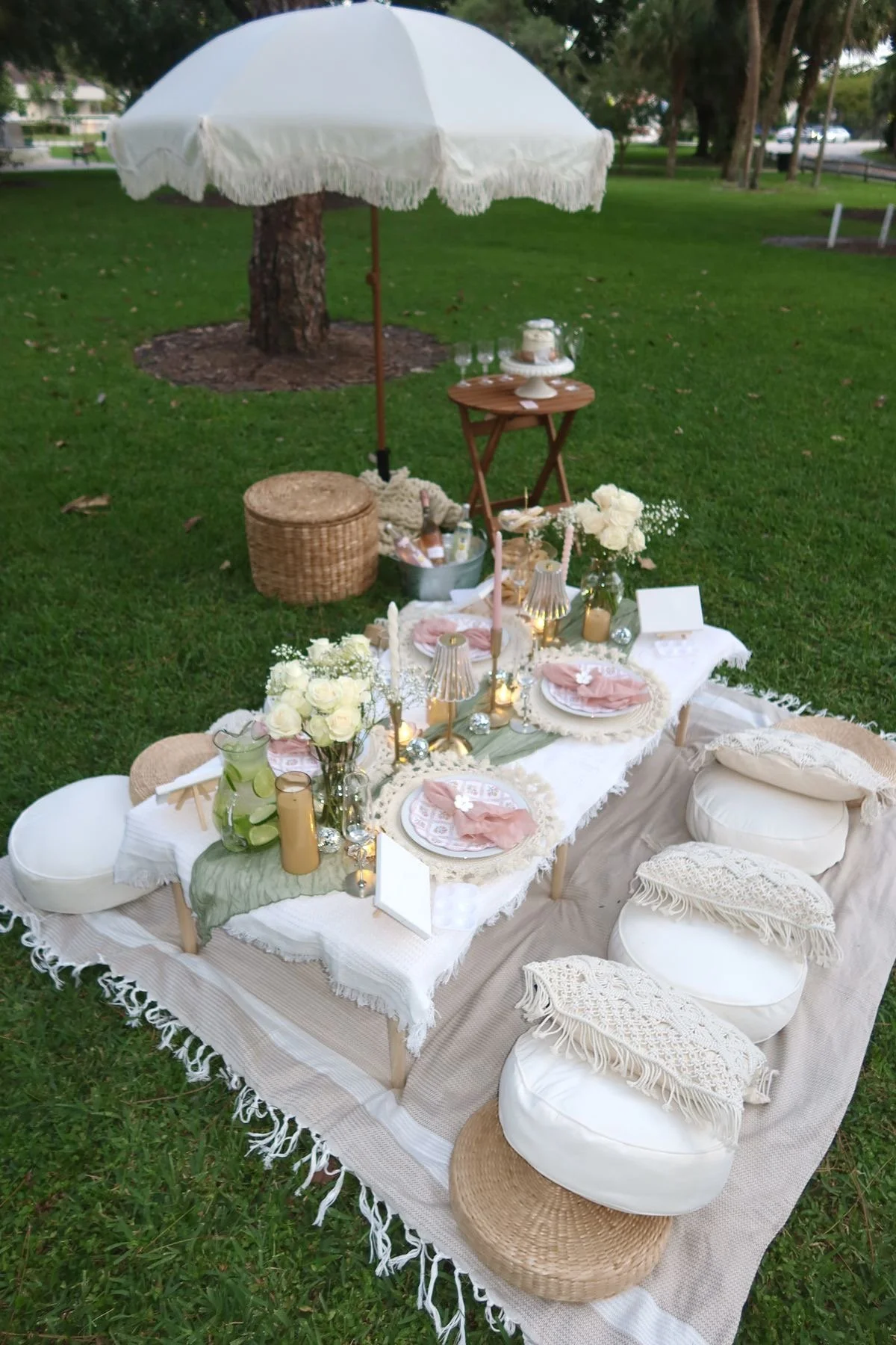 A picnic or outdoor dining setup with a long table on a beige and white woven rug on grass. The table has light pink napkins, floral plates, candles, vases with white flowers, and small lamps. Cushions and small poufs surround the table, and a large white umbrella shades part of it. In the background, there are trees and parked cars.