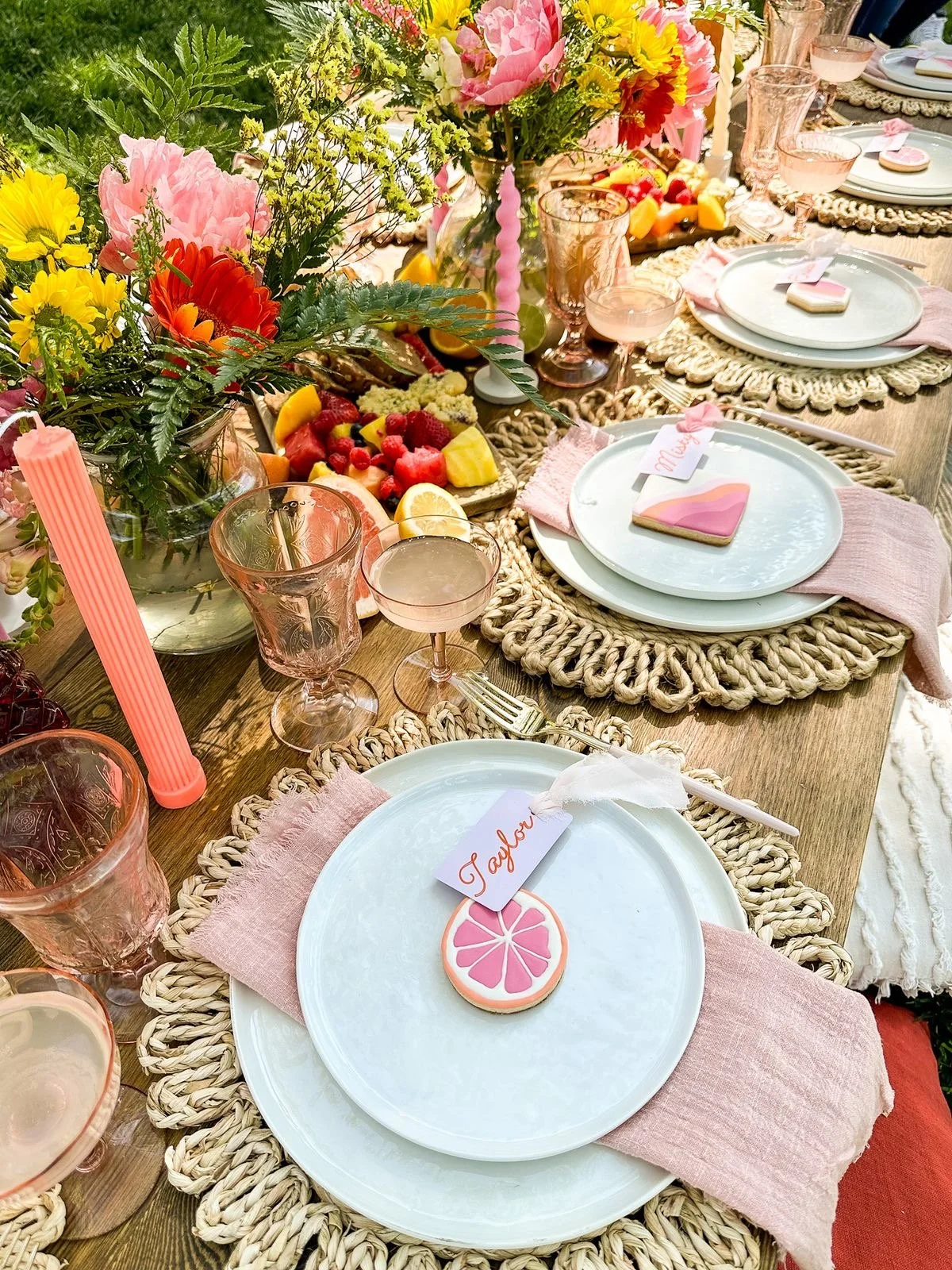 A table set for a celebration with white plates, pink cloth napkins, and cookies decorated with pink and orange slices and names like Taylor and Margot. The table features woven placemats, pink glassware, and a large bouquet of colorful flowers including pink, yellow, and red blooms, along with fresh fruit and pink candles.