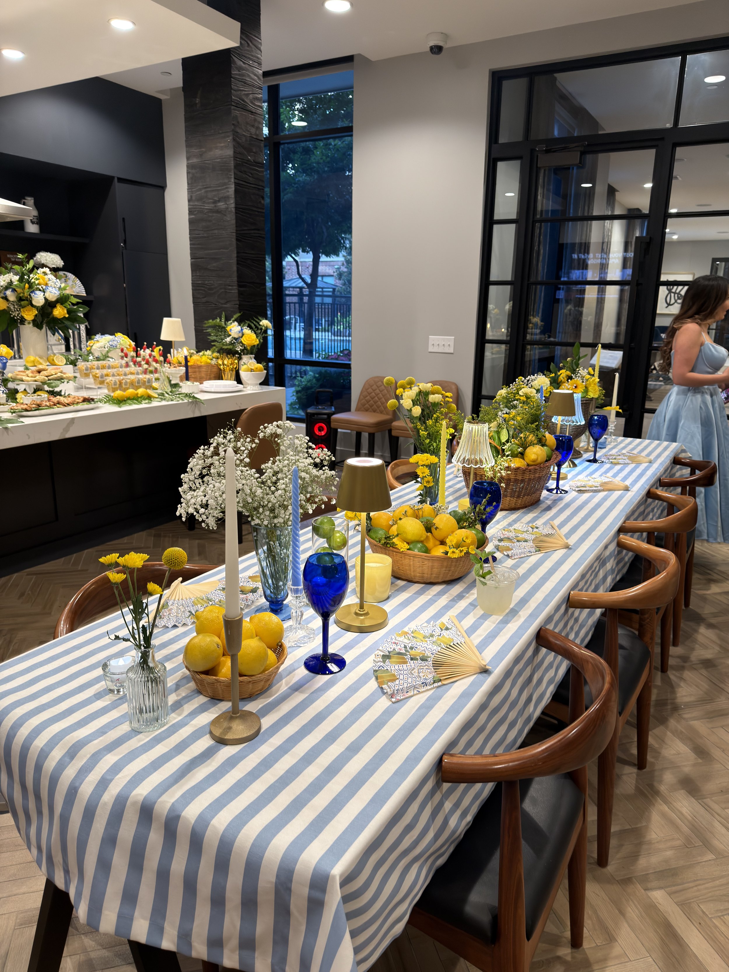A dining room decorated with flowers, lemons, and blue glassware on a table with a blue and white striped tablecloth. In the background, a buffet table displays various foods, flowers, and decorations. The room has large windows and modern black-framed glass doors, with a woman in a light blue dress standing to the right.