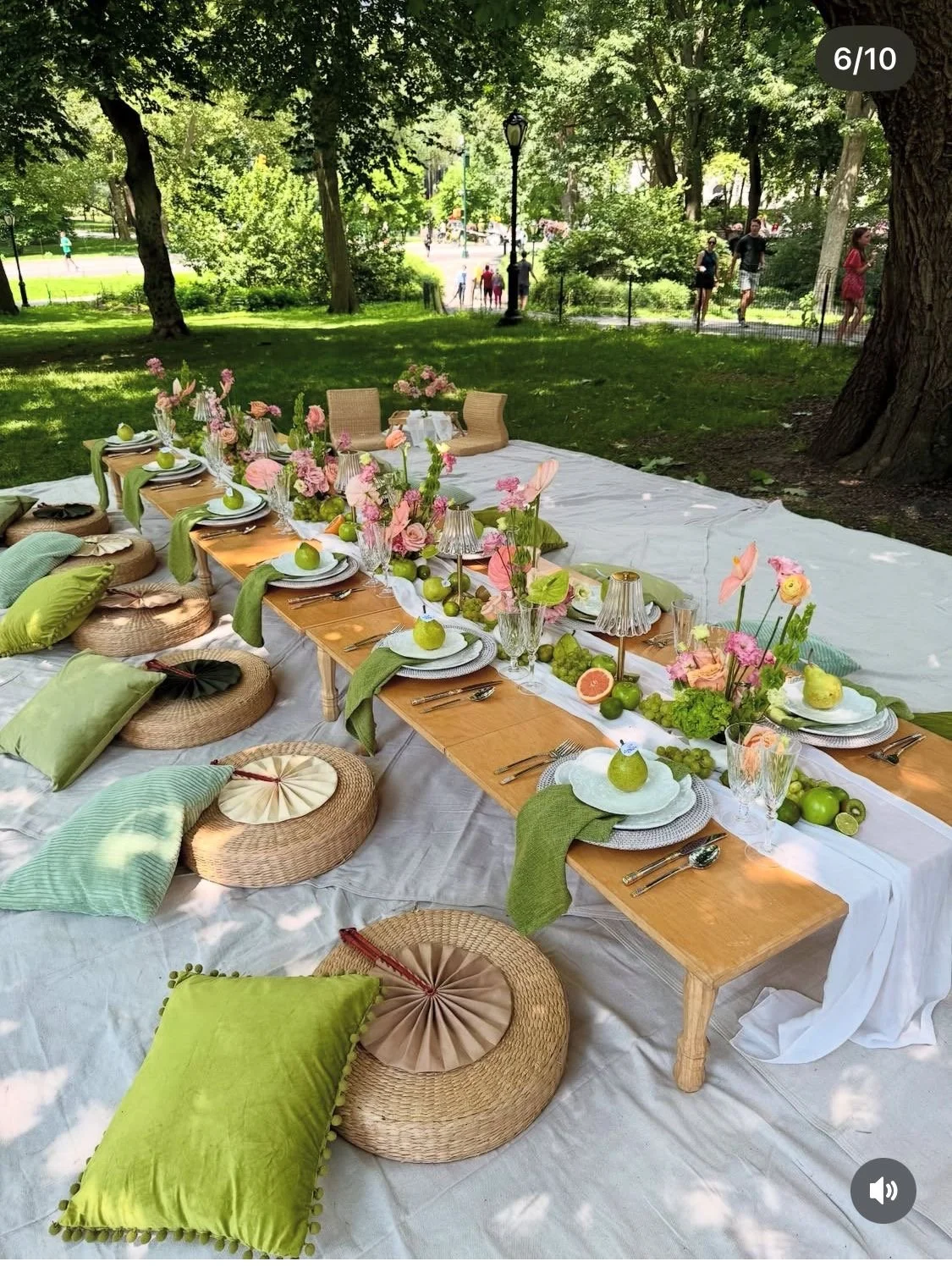 A picnic table set for an outdoor meal in a park, decorated with pink flowers, green apples, and glassware, with cushions and pillows on the ground around the table.