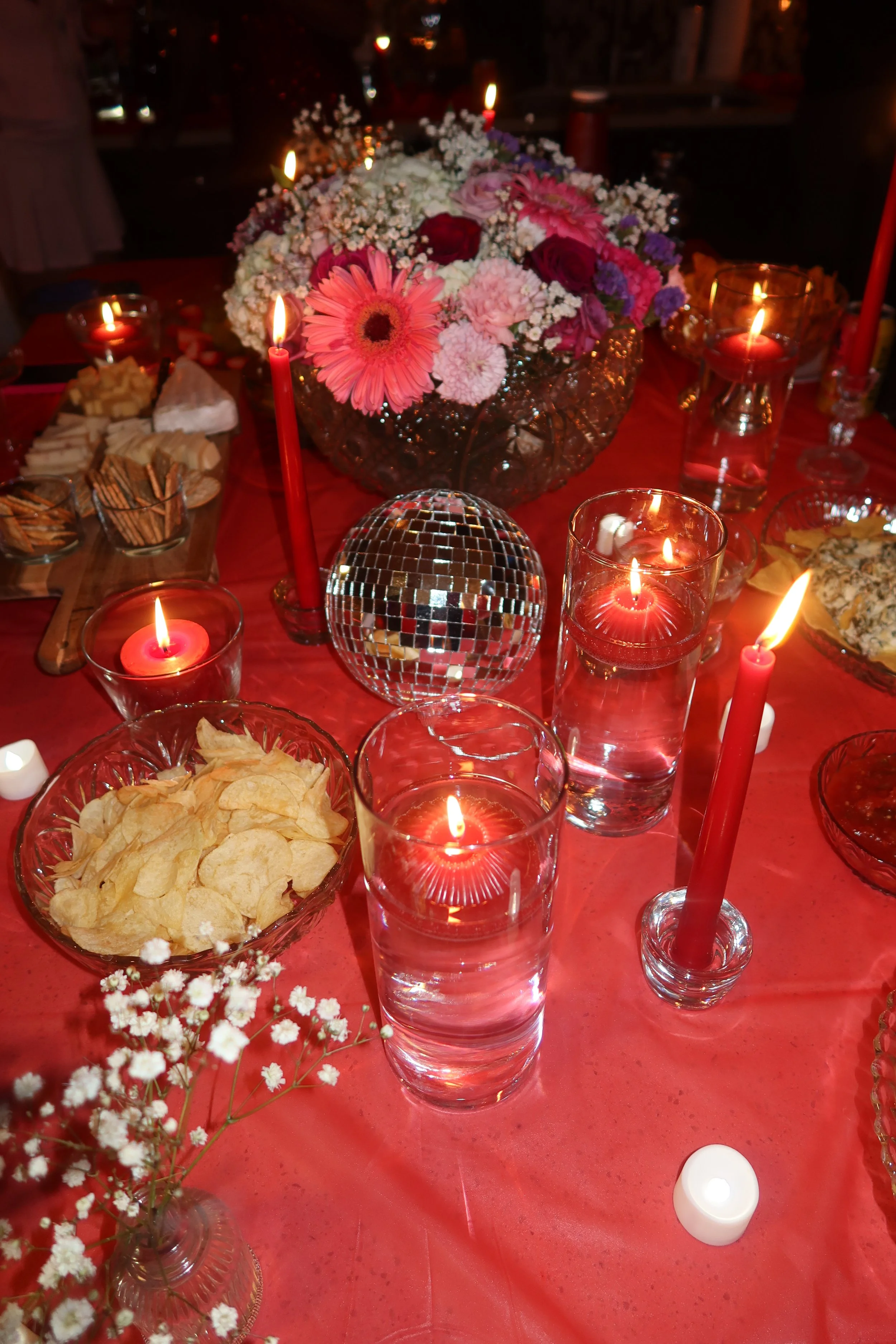A table decorated with pink and purple flowers in a glass bowl, surrounded by lit red candles in glass holders and a clear glass disco ball, with bowls of chips and snacks on a red tablecloth.