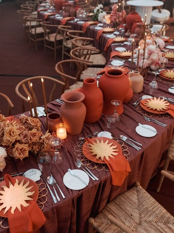 Decorated dining table set for a formal event with terracotta vases, candle, flowers, and orange napkins with a sun-shaped design.