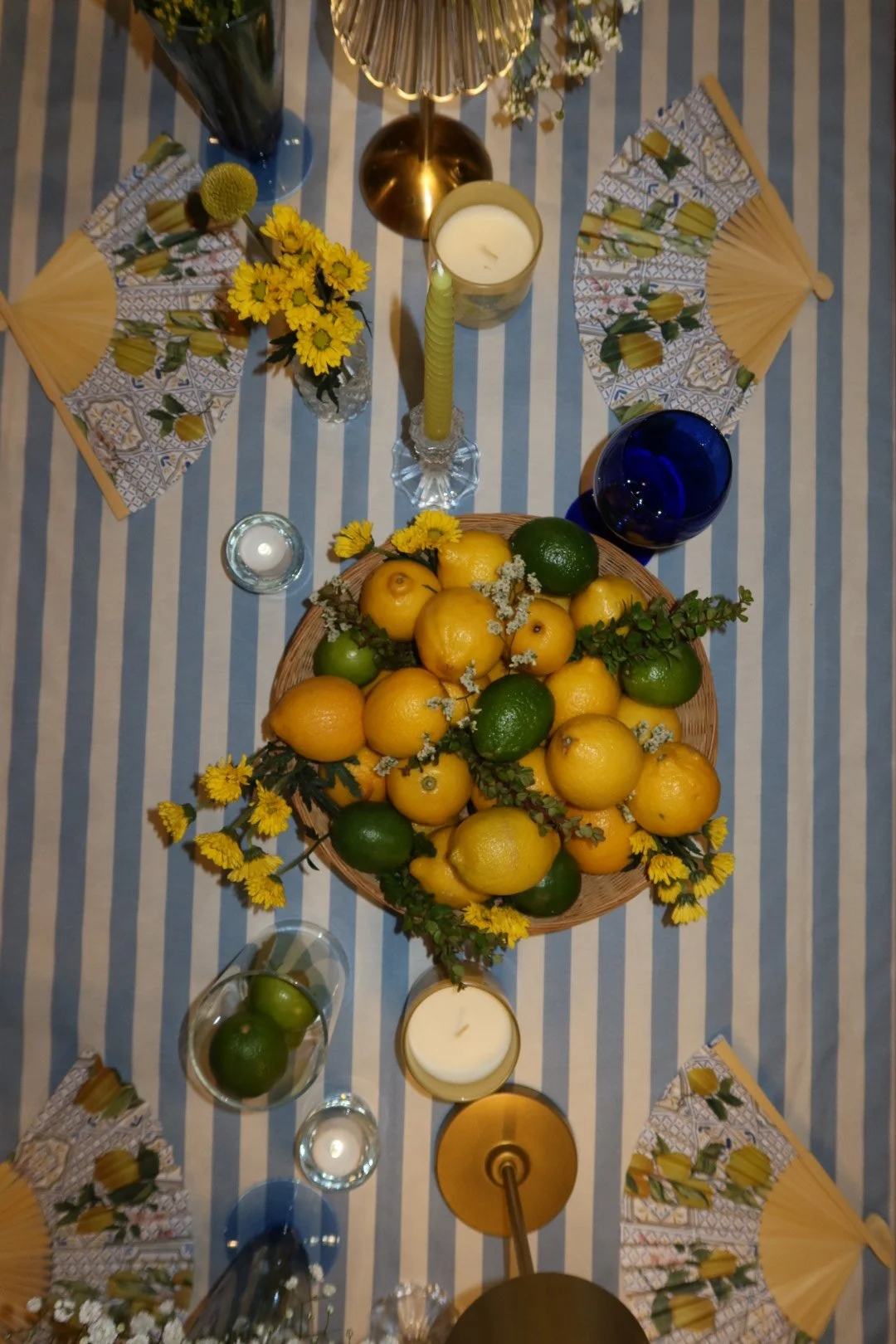 Top view of a dining table with a blue and white striped tablecloth, a basket full of lemons and limes, yellow flowers, candles, and decorative fans.