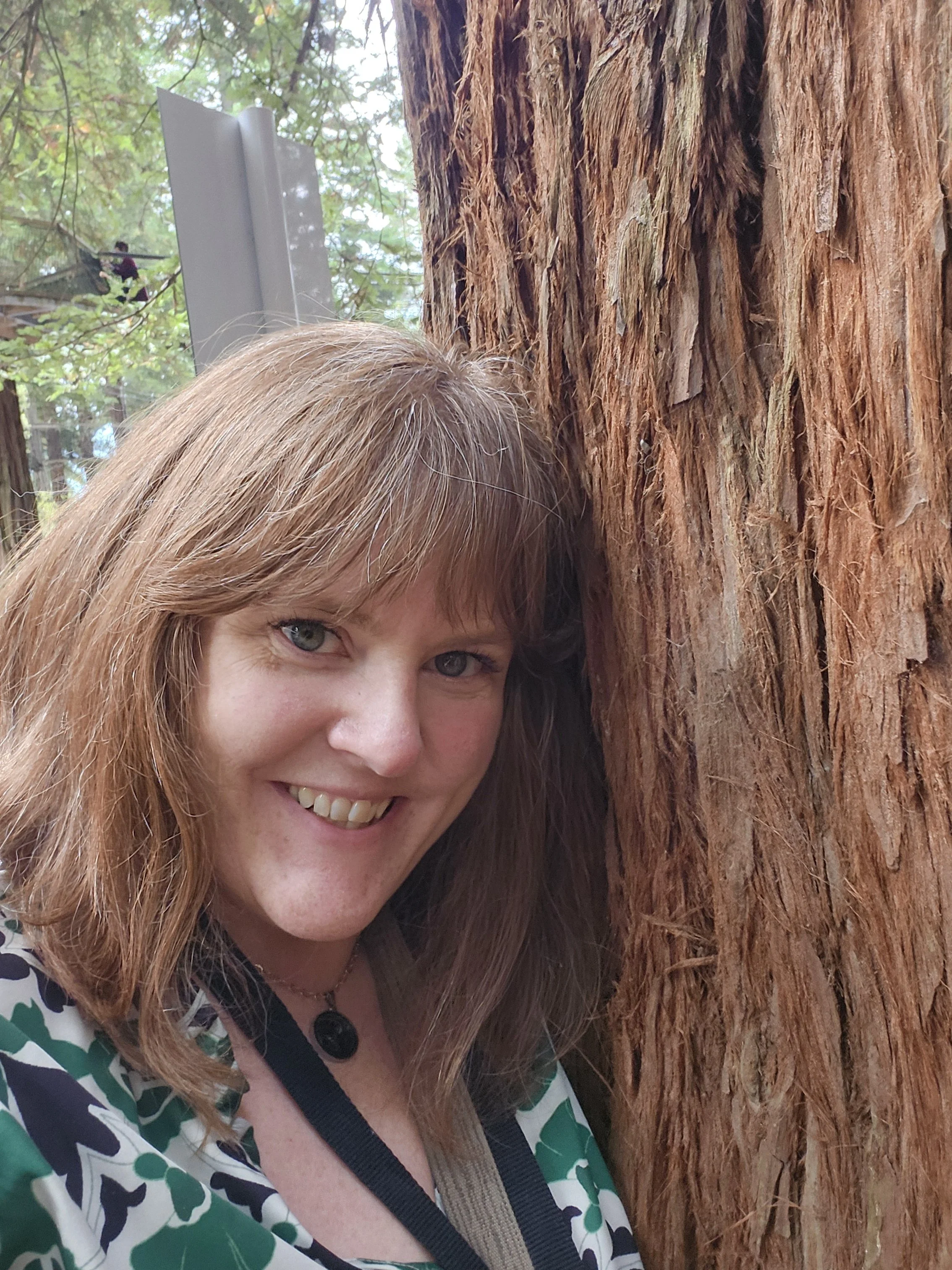 A woman with long red hair and blue eyes smiling, standing next to a large tree trunk with textured, reddish-brown bark, outdoors with green leaves and branches in the background.