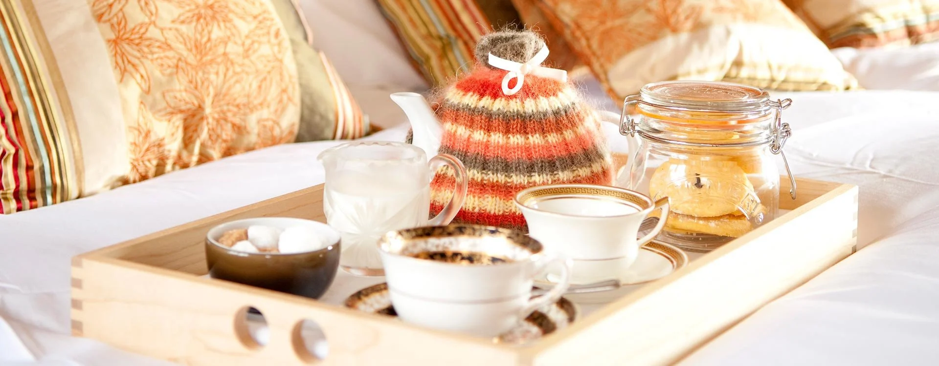 A breakfast tray with cups of coffee, a bowl of marshmallows, a teapot, a jar of cookies, and a cozy striped knit tea cozy, placed on a bed with pillows.