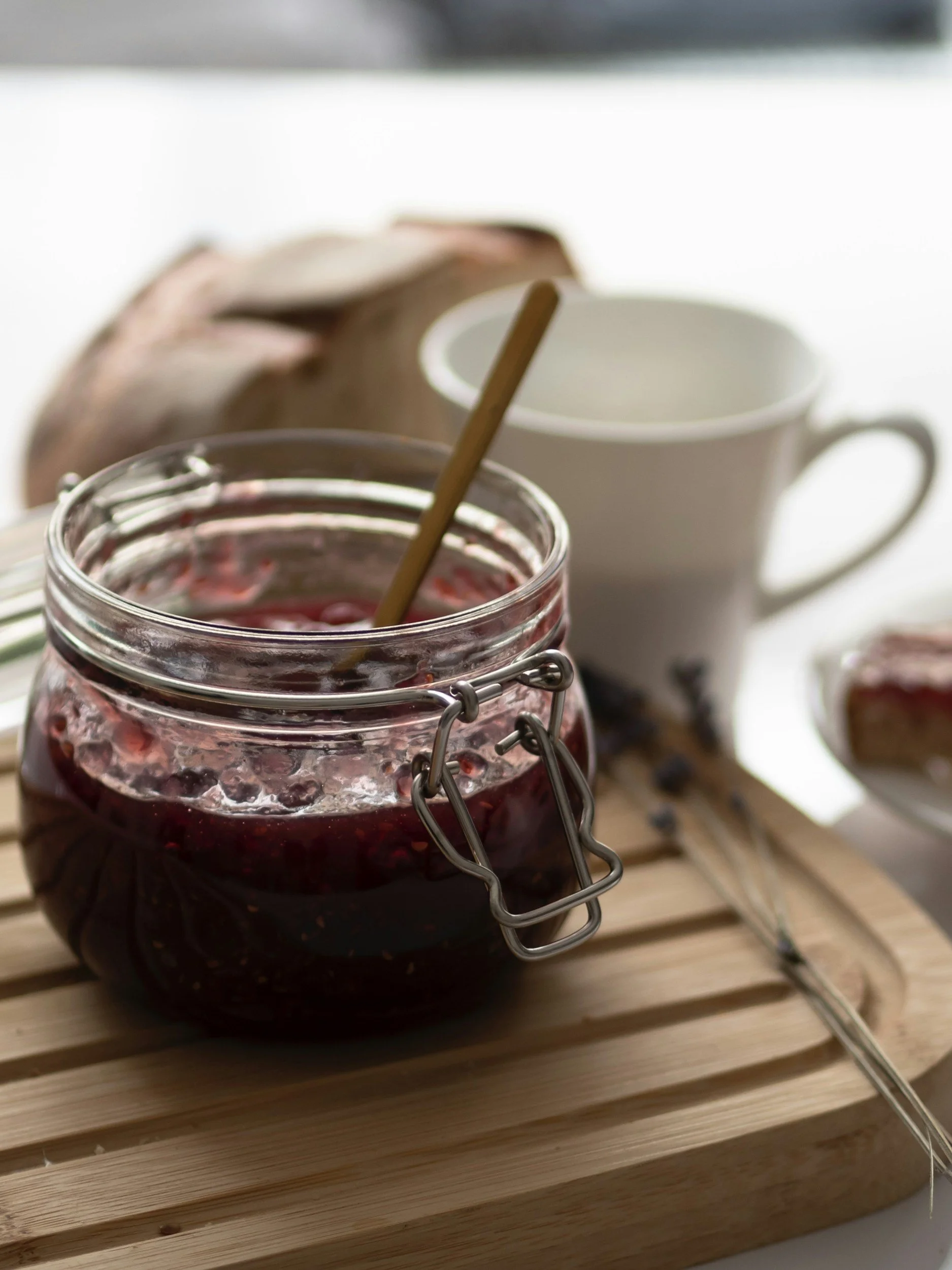 A glass jar filled with red jam or jelly, with a wooden spoon inside. In the background, there are two white mugs, on a wooden surface.