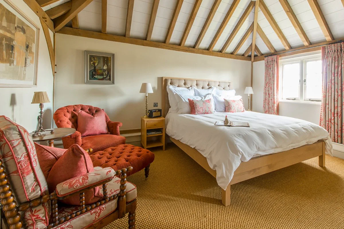 A cozy bedroom with a high, vaulted ceiling featuring exposed wooden beams, a large bed with white linens and pink decorative pillows, flanked by two nightstands with lamps, and a sitting area with a red upholstered armchair and matching ottoman, all illuminated by natural light from a window with floral curtains.