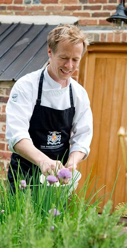 A chef wearing a white coat and black apron smiling and tending to purple flowers and green plants in a garden.
