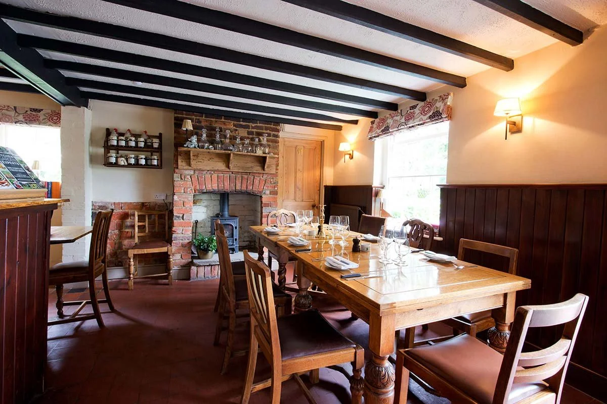 A cozy dining room with a wooden table set for a meal, surrounded by wooden chairs, a fireplace, and brick accents. The room has large windows with floral valances and warm wall-mounted lights.