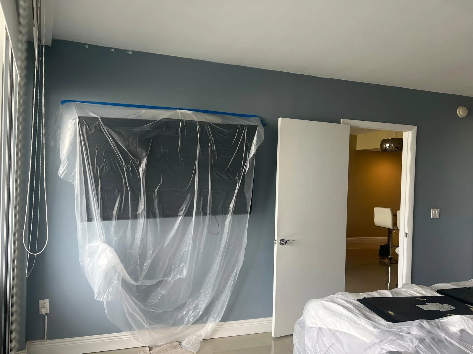 A room with light blue walls, a mounted flat-screen TV covered with plastic, an open white door leading to a room with a yellow wall and a white chair, and a bed with protective plastic.