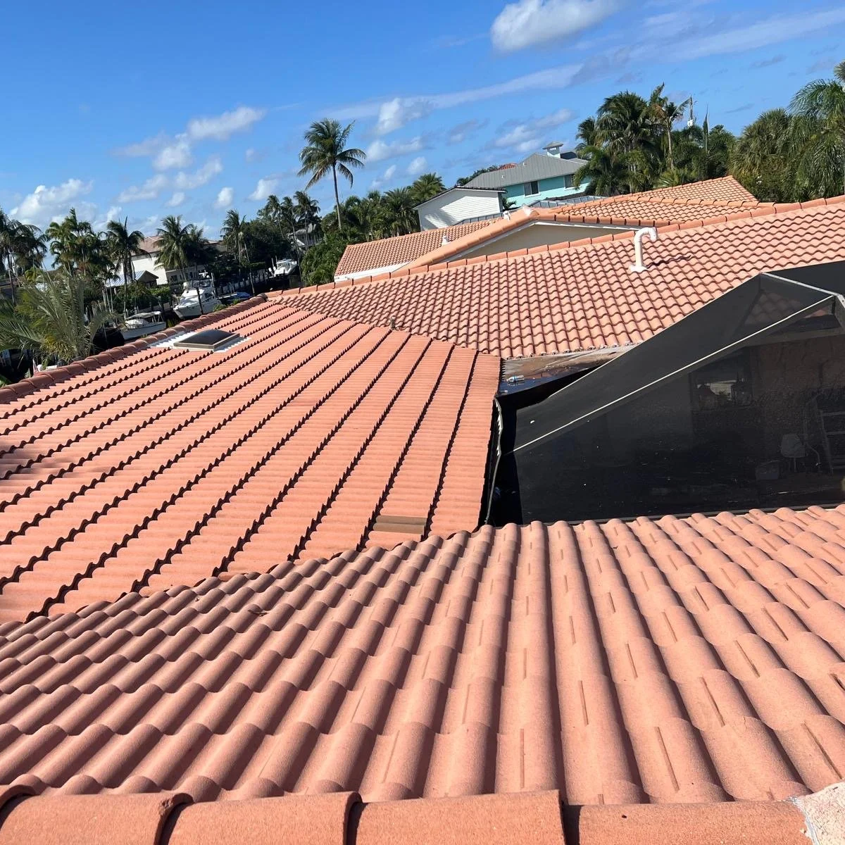 Aerial view of tiled rooftops in a tropical neighborhood with palm trees and modern houses under a blue sky with scattered clouds.