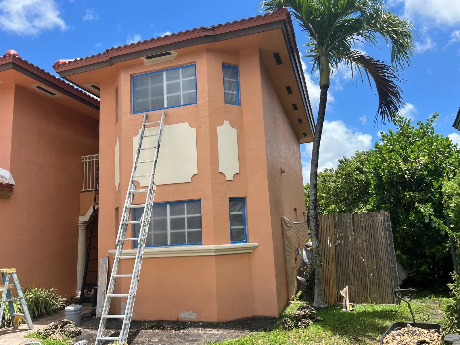 A three-story house with peach-colored stucco walls, blue plastic-covered windows, and a red tiled roof. A tall aluminum ladder leans against the house, with a worker visible near a side gate. The yard has lush green trees, including a tall palm, and a wooden fence. The sky is partly cloudy with patches of blue.