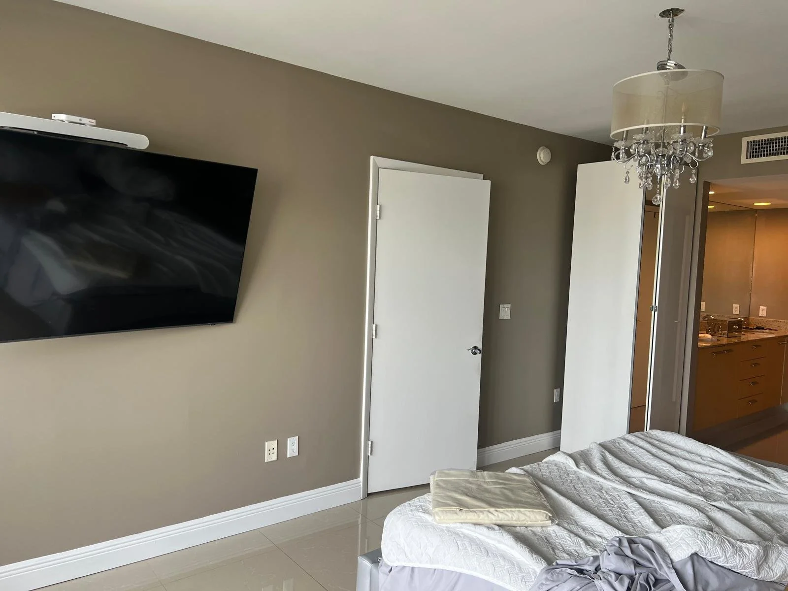 A bedroom with a mounted flat-screen TV, a white door slightly open, a bed with a white textured blanket, a beige pillow, and a folded gray piece of clothing. There is a crystal chandelier hanging from the ceiling and a closet with sliding doors in the background.