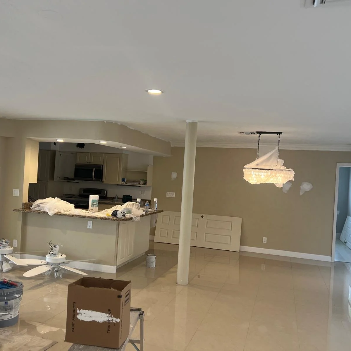 Living room and kitchen under renovation with tools, supplies, and protective coverings on the ceiling and chandelier.
