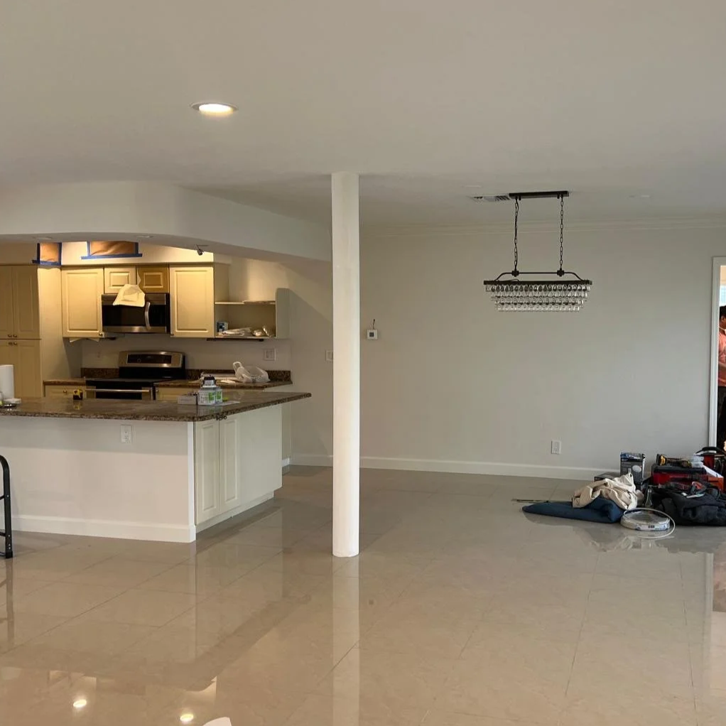 Empty kitchen and living area with some tools and materials on the floor, a chandelier hanging from the ceiling, and a partial view of a person near a doorway.