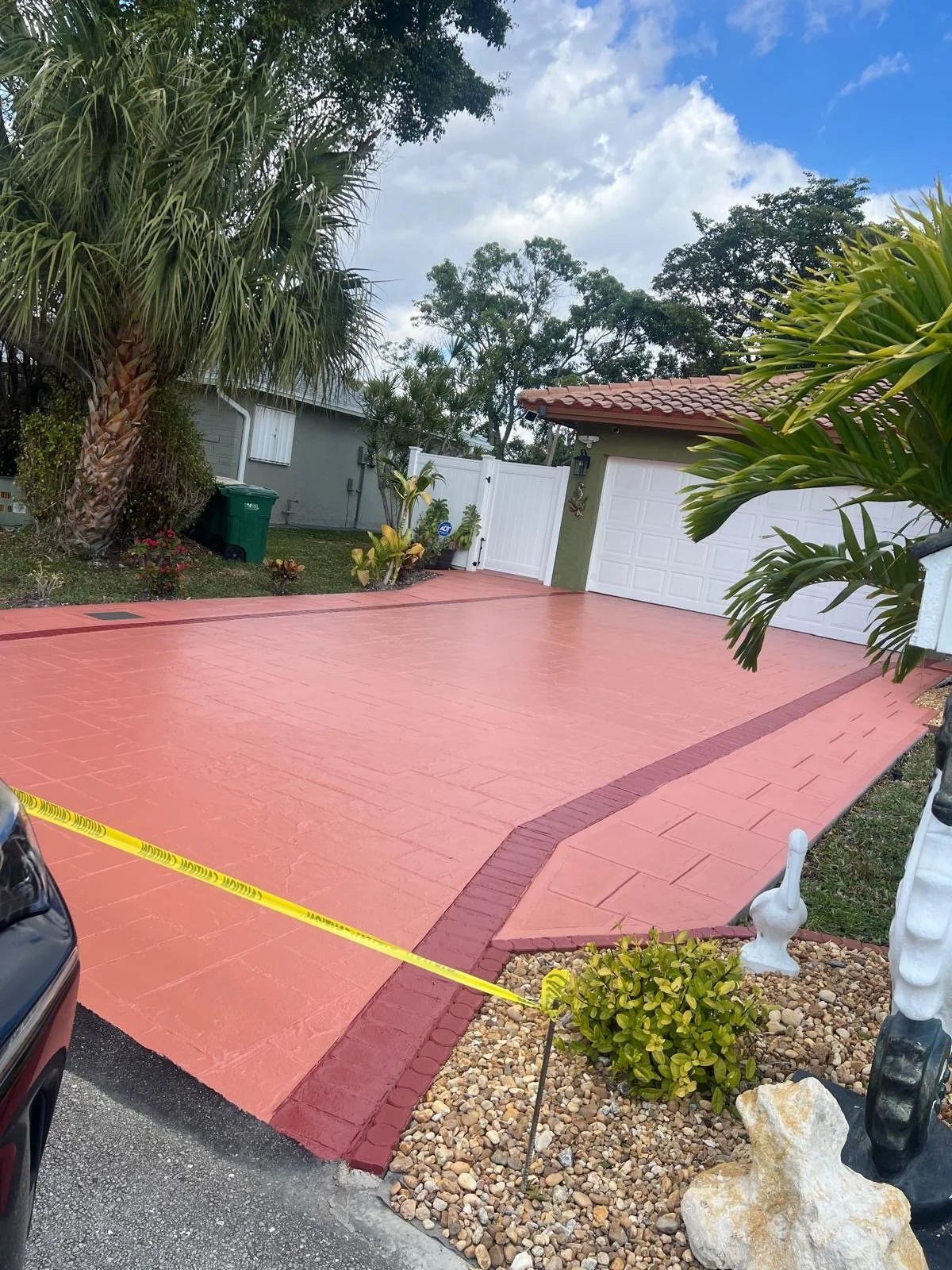 A backyard with a red concrete patio, white fence, and a garage with white doors. There are large palm trees and other plants around the yard, and a yellow caution tape in the foreground.