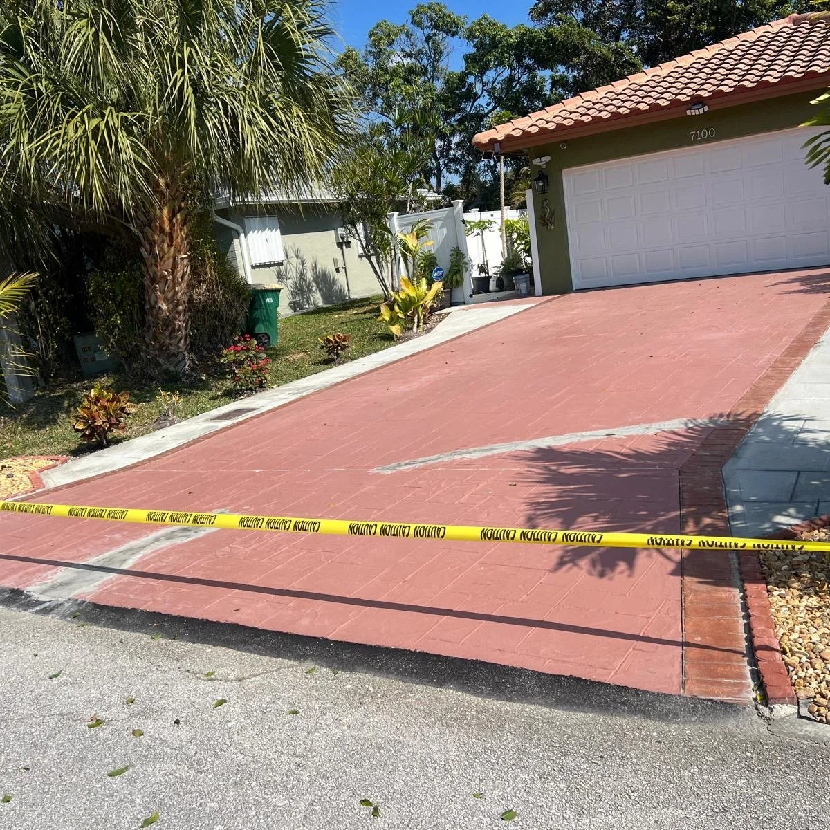 Driveway with yellow caution tape blocked off, leading to a house with a white garage door and a tiled roof, surrounded by tropical plants and trees.