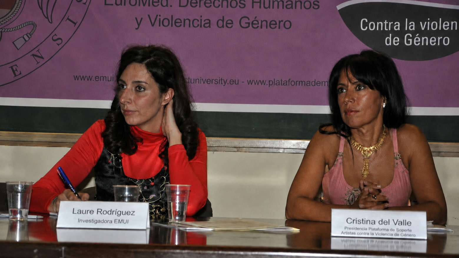 Dos mujeres sentadas en una mesa durante una conferencia o evento. La mujer a la izquierda lleva una blusa roja y tiene el cabello rizado oscuro, con un cartel que dice 'Laure Rodríguez, Investigadora EMUI'. La mujer a la derecha lleva una blusa rosa y tiene el cabello negro y liso, con un cartel que dice 'Cristina del Valle, Presidencia Plataforma de Soporte Artistas contra la Violencia de Género'.