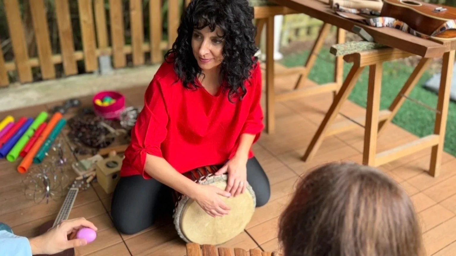 Mujer con cabello rizado y ropa roja tocando un tambor mientras está sentada en un suelo de madera, rodeada de instrumentos musicales y niños en un ambiente al aire libre.