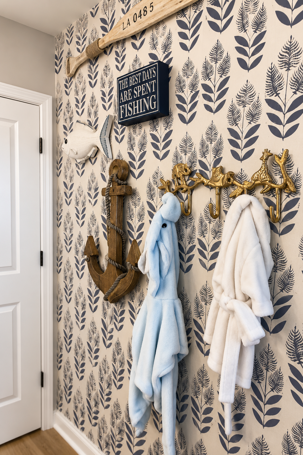 Decorative wall with nautical theme, featuring a fish wall sculpture, a wooden anchor, a mounted sign that reads 'The best days are spent fishing,' and hooks with plush bathrobes hanging on a gold hook rack shaped like animals.
