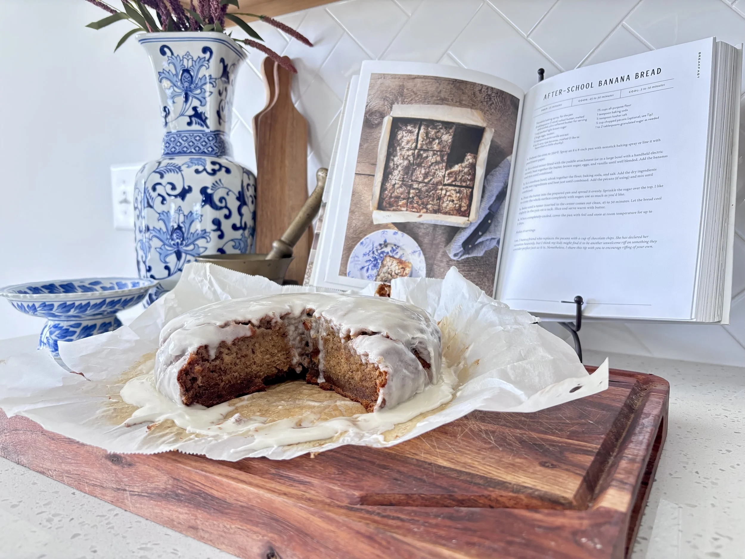 A partially sliced banana bread with frosting on a wooden cutting board, with a blue and white floral vase, a small bowl, and a cookbook open showing a banana bread recipe in the background.