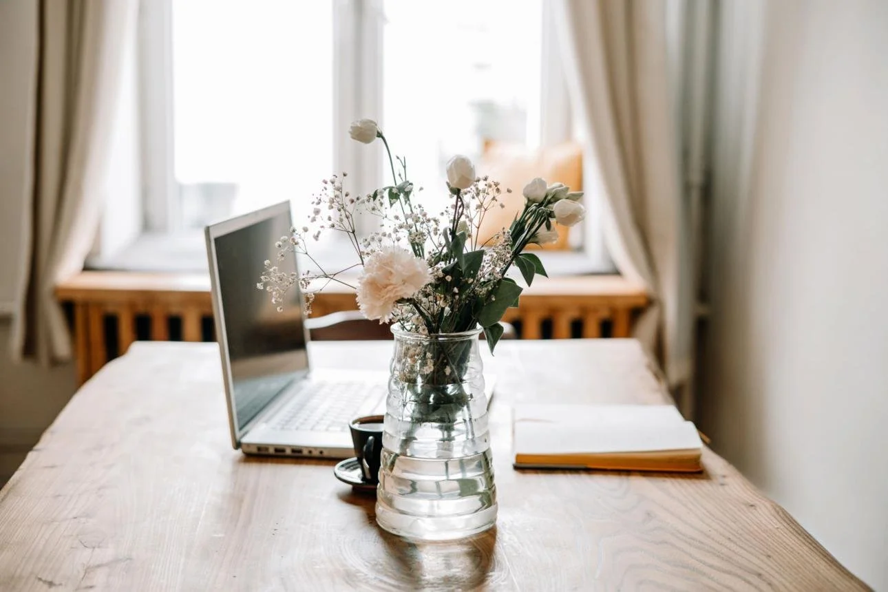 A wooden table with a laptop, a closed notebook, and a glass vase with white flowers against a window with curtains.