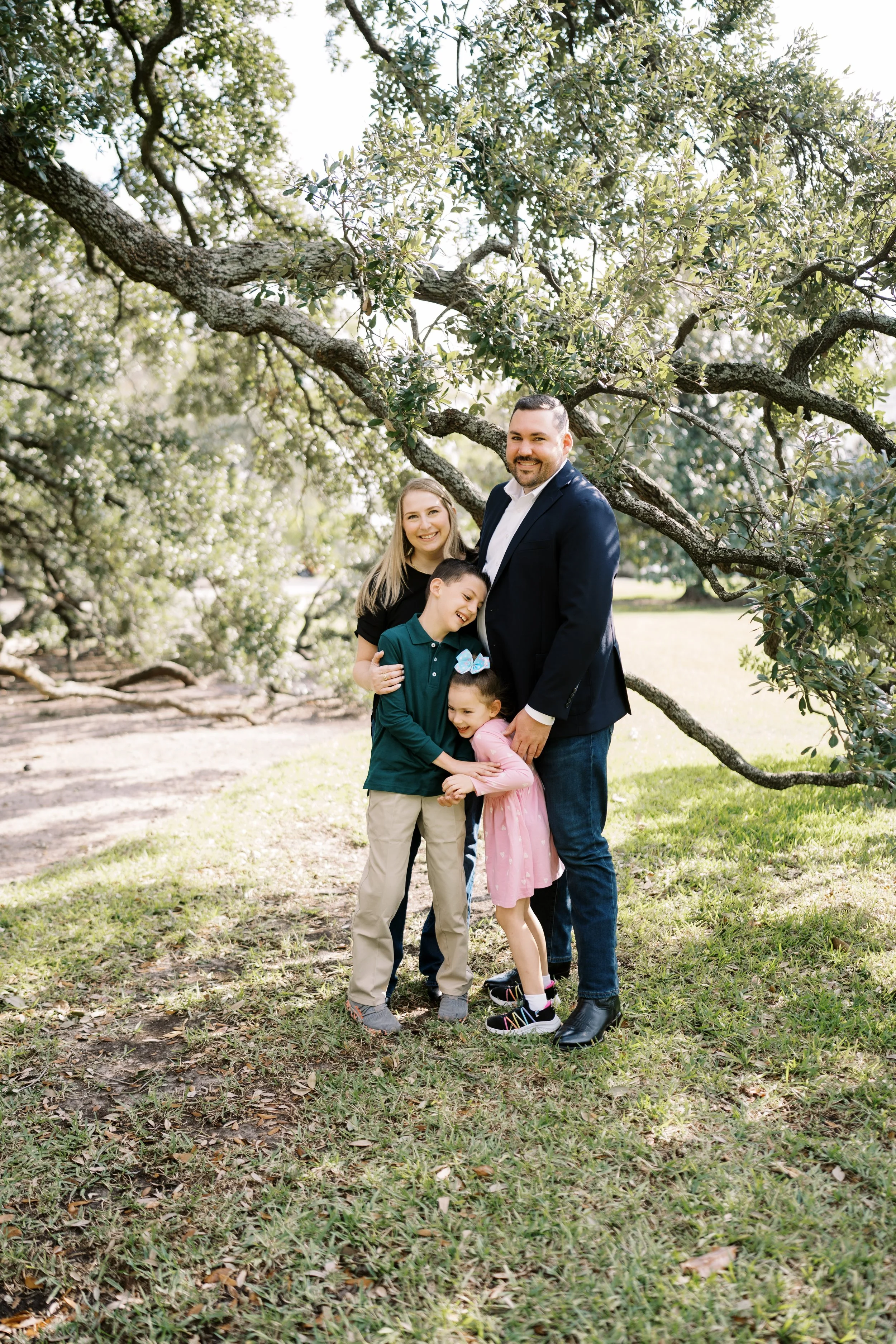 A family of five standing under a large tree in a park, smiling for the camera. The family includes two adults and two children, with the children hugging each other.