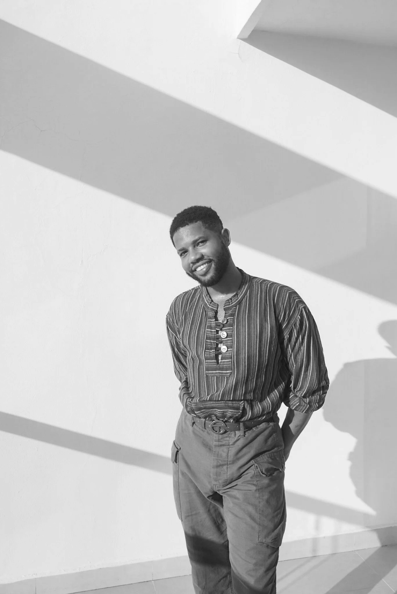 A smiling man with a beard and short curly hair standing against a white wall with dramatic shadows cast from above. He is wearing a striped shirt, khaki cargo pants, and a belt. His right hand is resting in his pocket and he is looking at the camera.