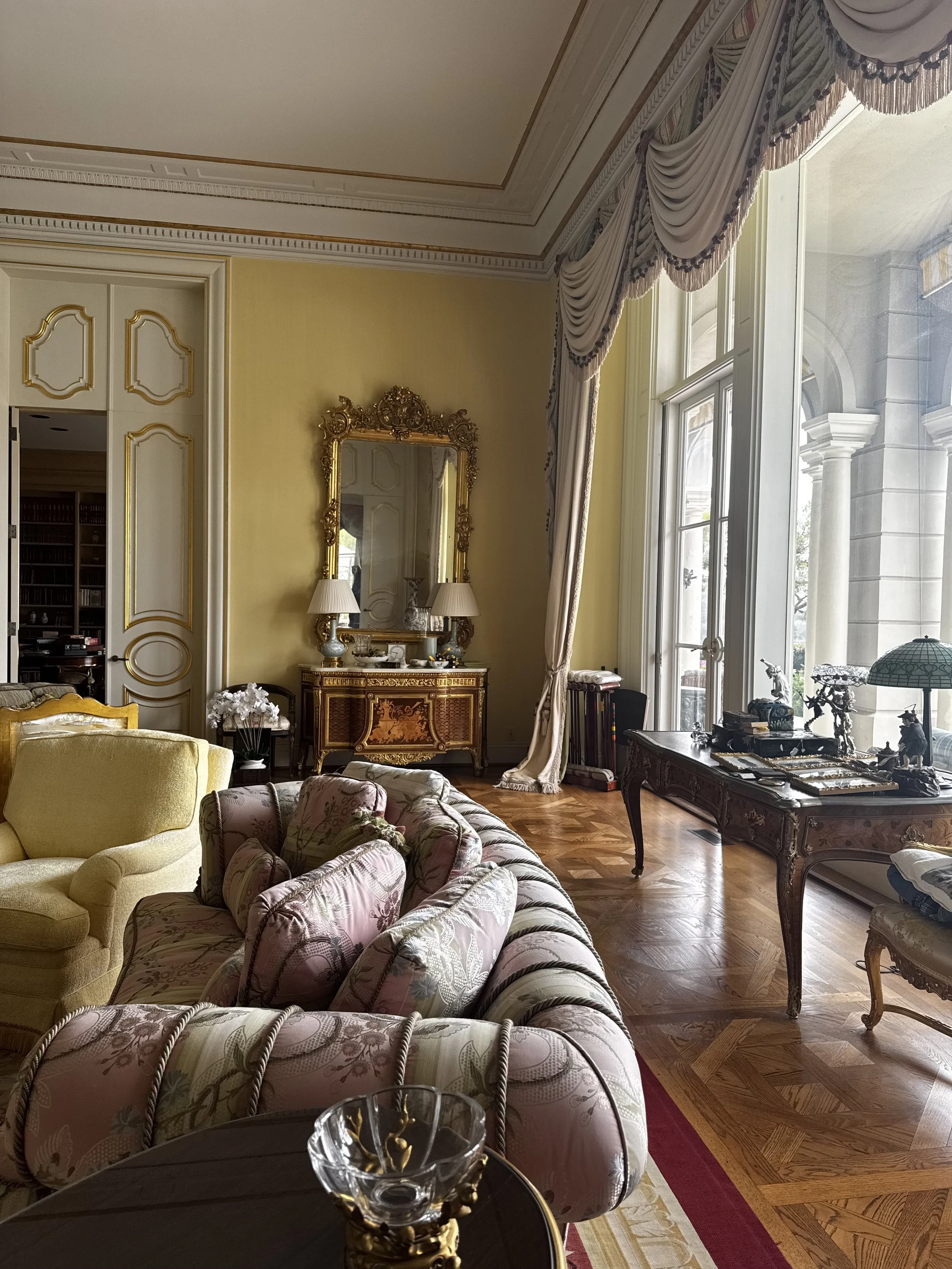 Elegant living room with high ceiling, large windows with ornate curtains, vintage furniture including floral and yellow upholstered sofas, a wooden floor with intricate parquet pattern, a gold-framed mirror, and decorative items on tables.