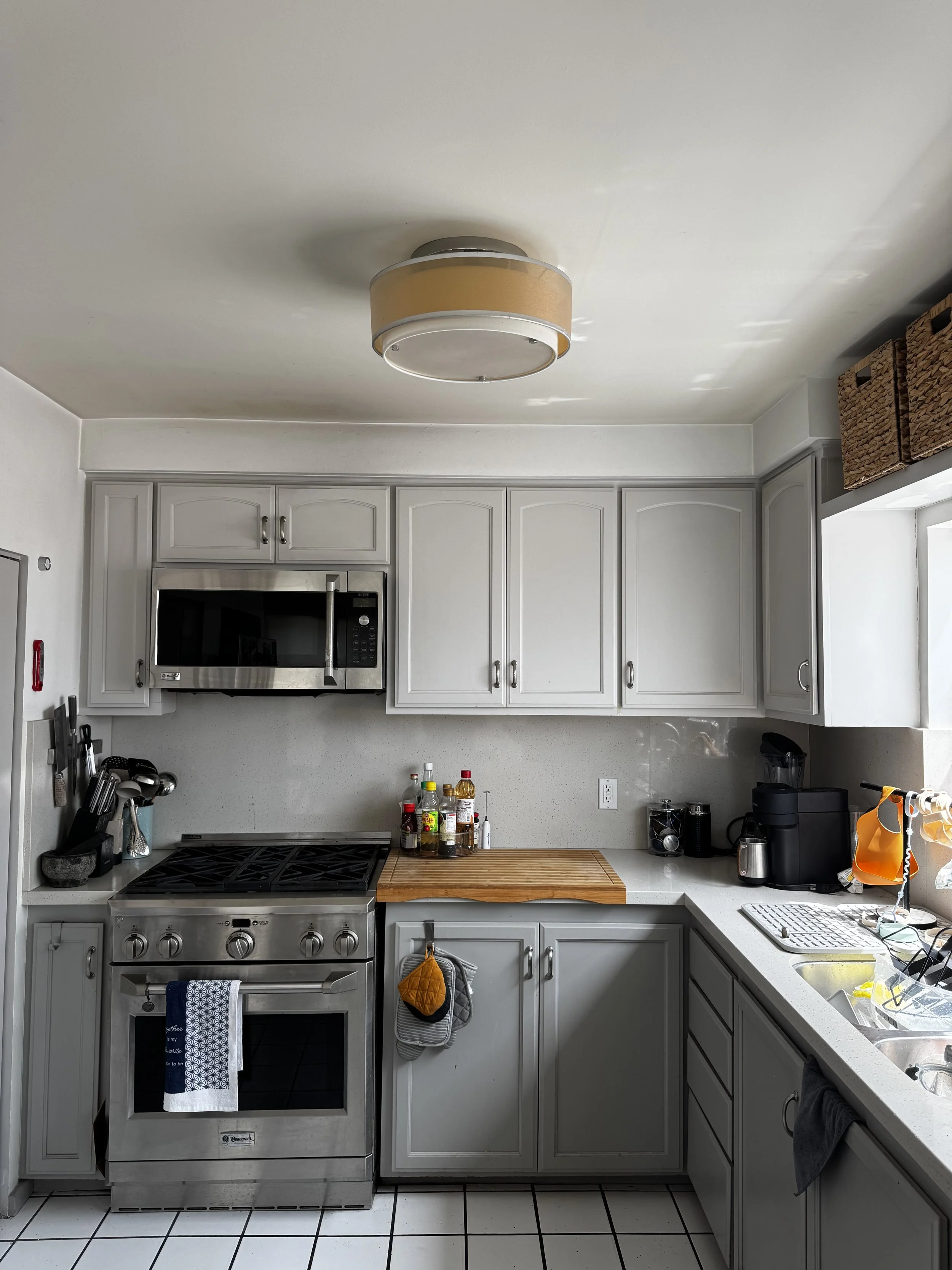 A kitchen with white cabinets, stainless steel oven and microwave, countertops with various condiment bottles, a cutting board, and a sink with dish drying rack. There are kitchen utensils on the wall, and a black coffee machine on the counter. The floor is tiled with white and black grout.
