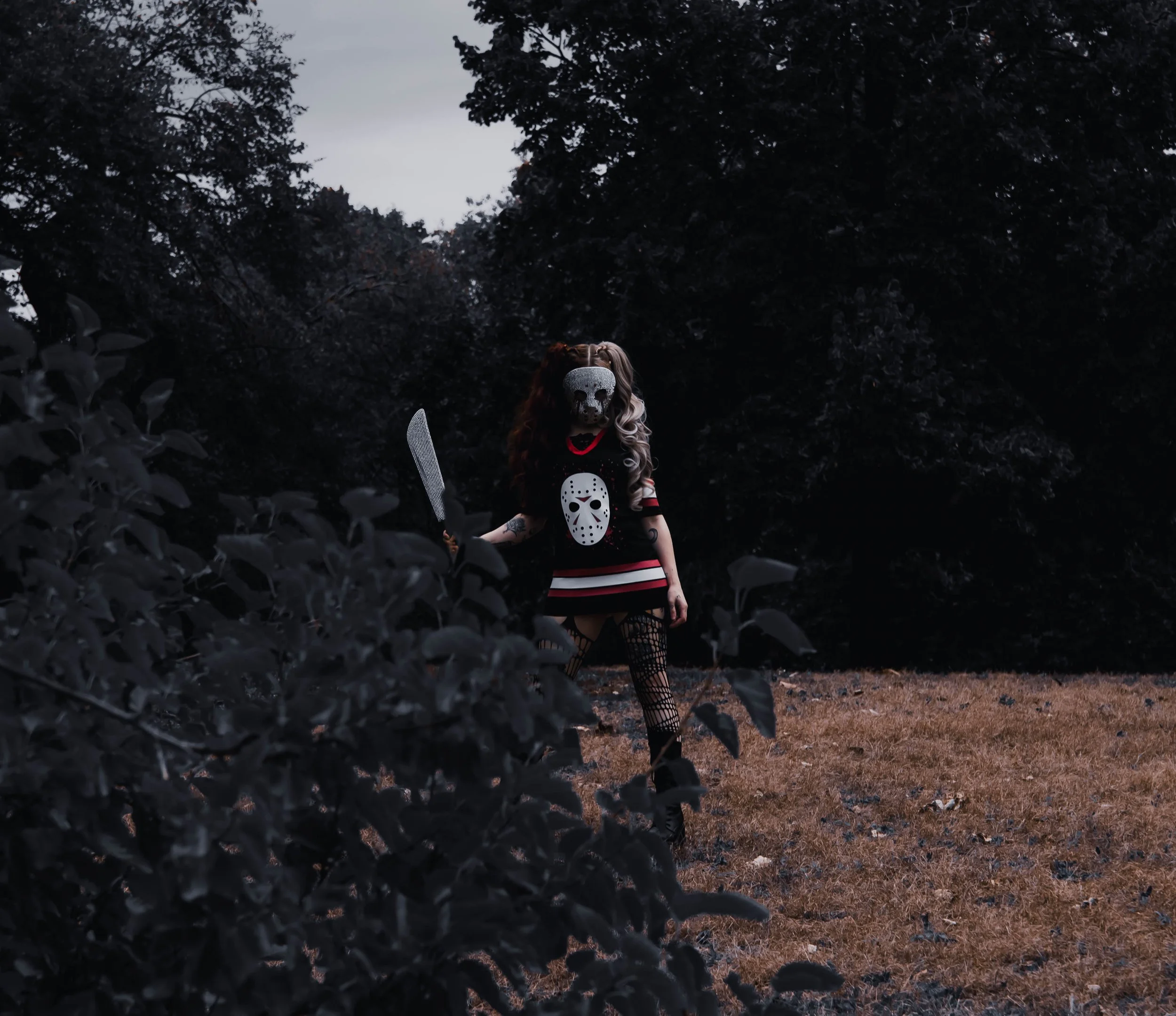 Person wearing a hockey mask stands in a wooded area during dusk, holding a large knife, with dark trees in the background.