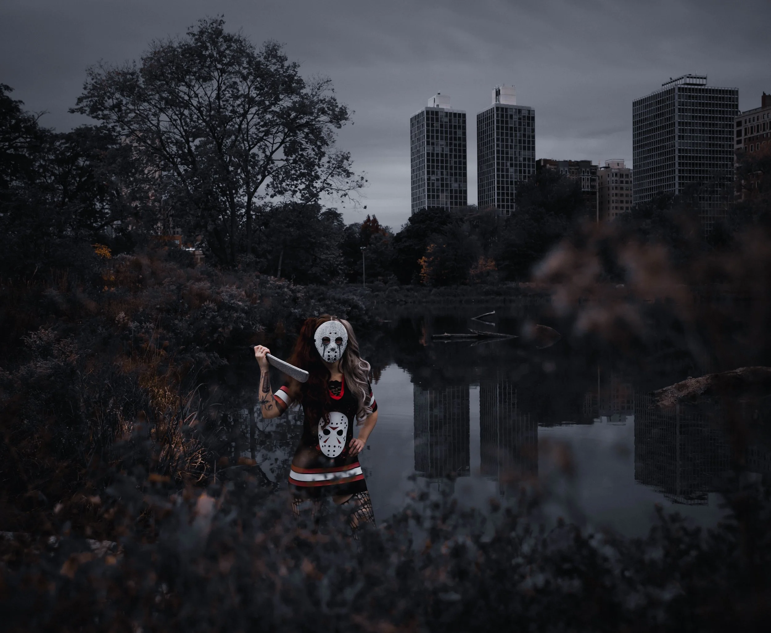A person dressed in a horror costume, wearing a hockey mask, and holding a weapon, standing near a body of water with a city skyline and multiple high-rise buildings in the background under an overcast sky.