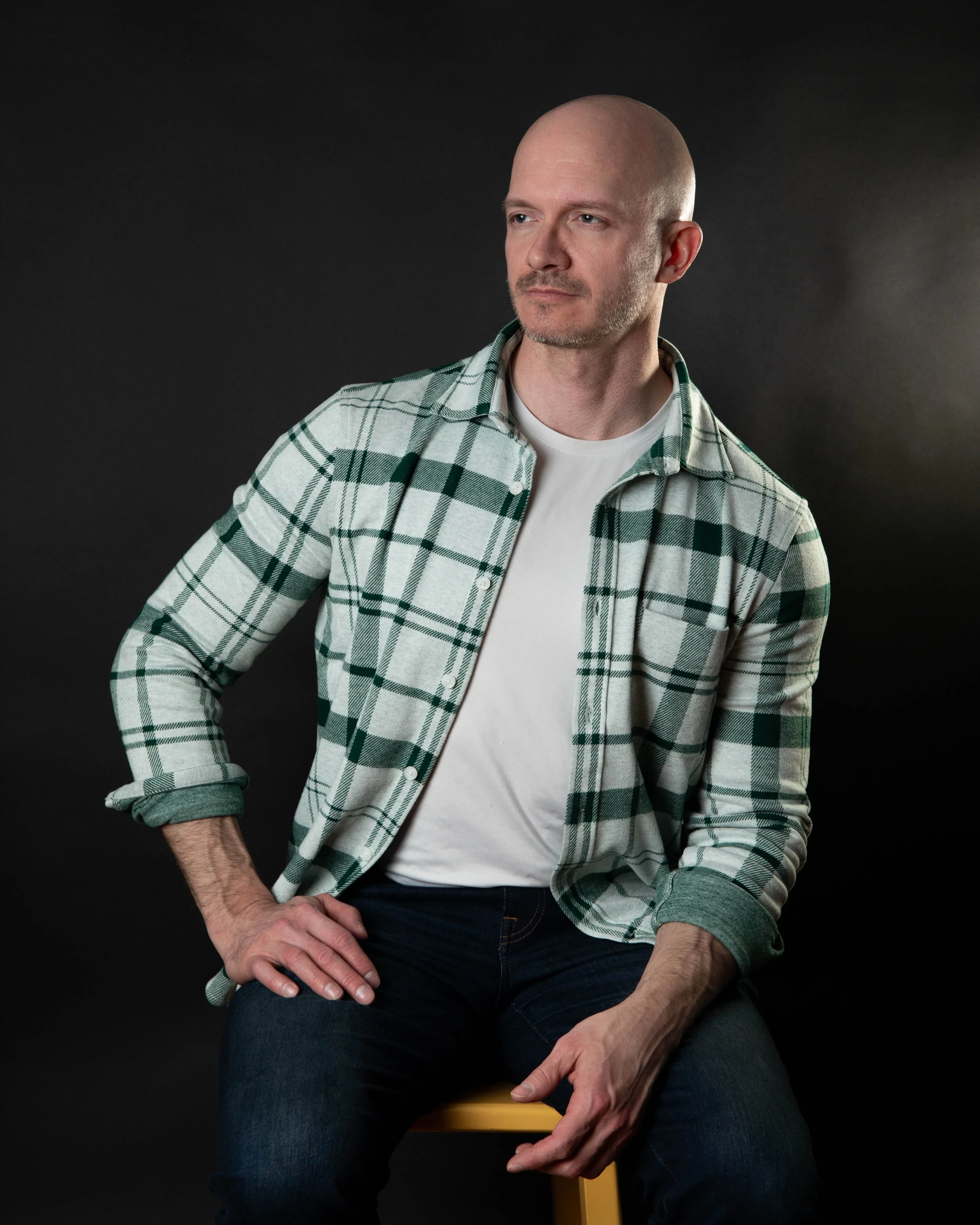 A bald man in a green and white plaid shirt and white t-shirt, sitting on a yellow stool against a black background, with one hand resting on his thigh.