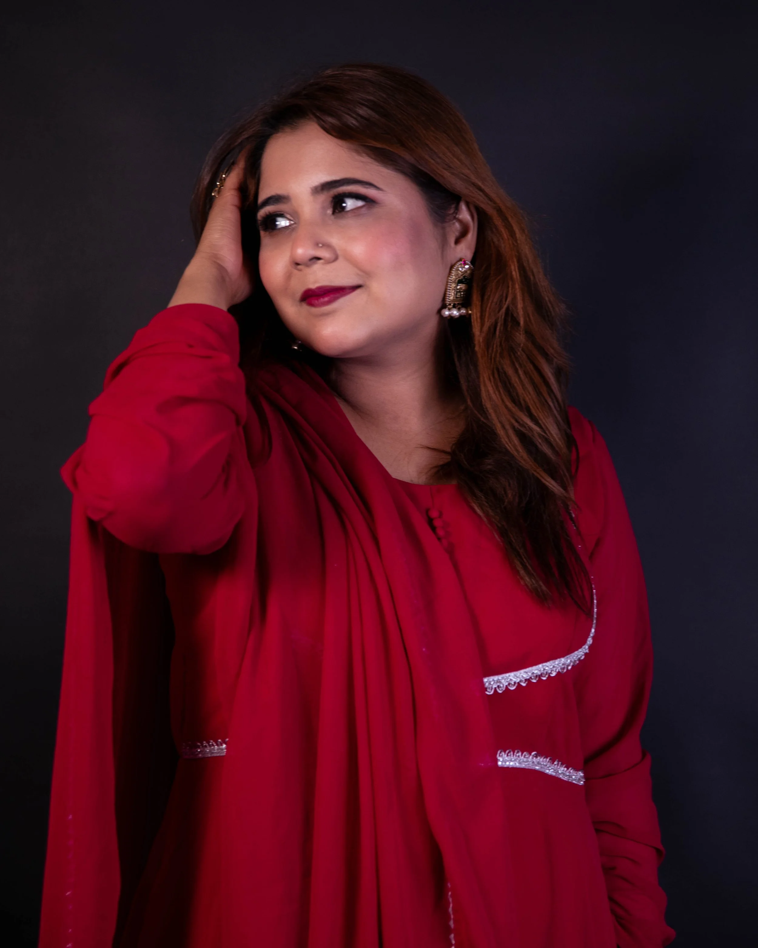 A woman with medium brown hair, wearing a red dress with lace sleeve accents, posed with her hand on her head against a dark background.