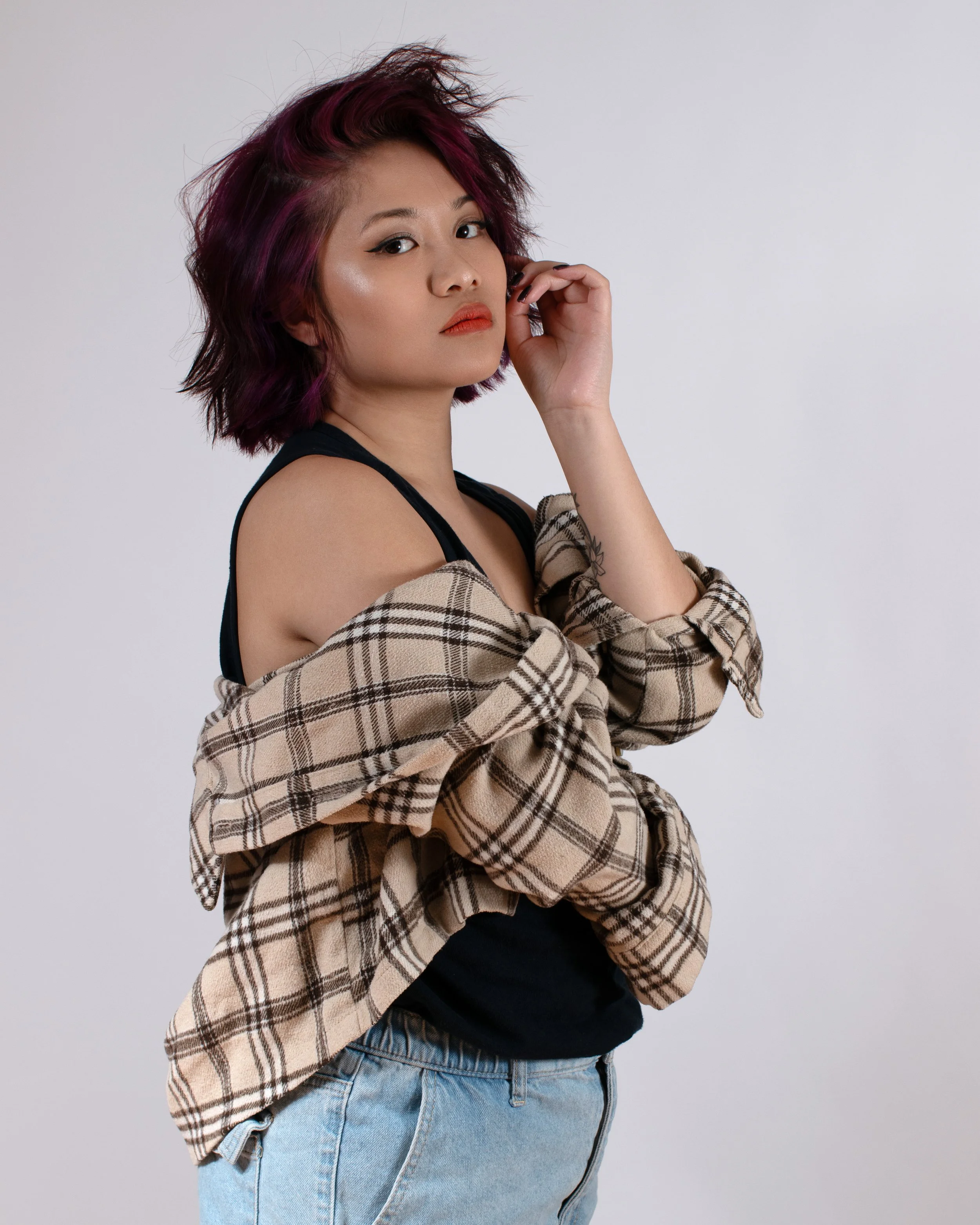 Young woman with short dark hair streaked with purple, wearing a black tank top, beige plaid shirt, and light blue jeans, posing with her hand near her face against a plain white background.