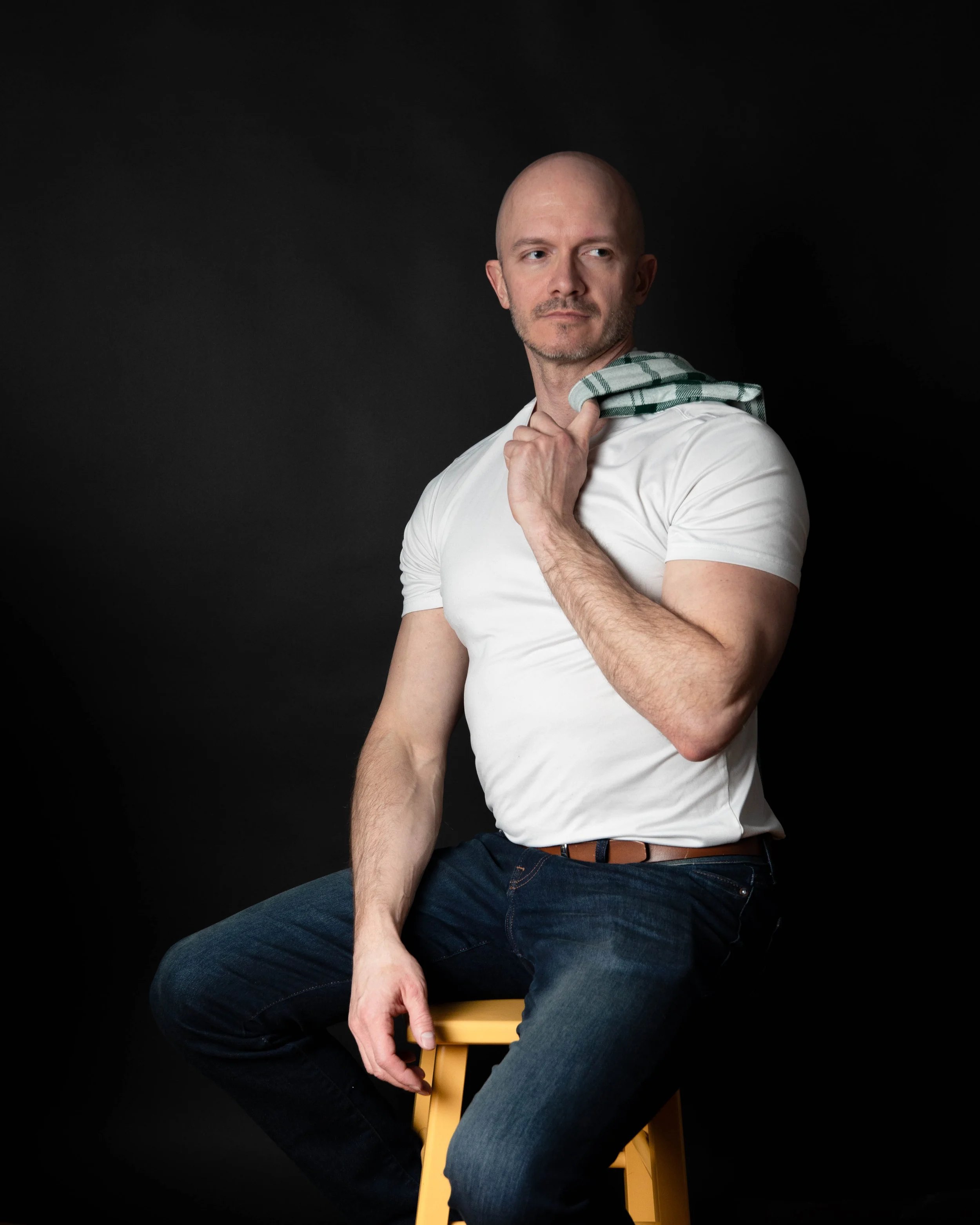 A bald man with a beard wearing a white T-shirt and dark jeans is sitting on a yellow wooden stool against a black background. He's holding a checkered shirt over his shoulder with his right hand, while his left arm rests on his knee.