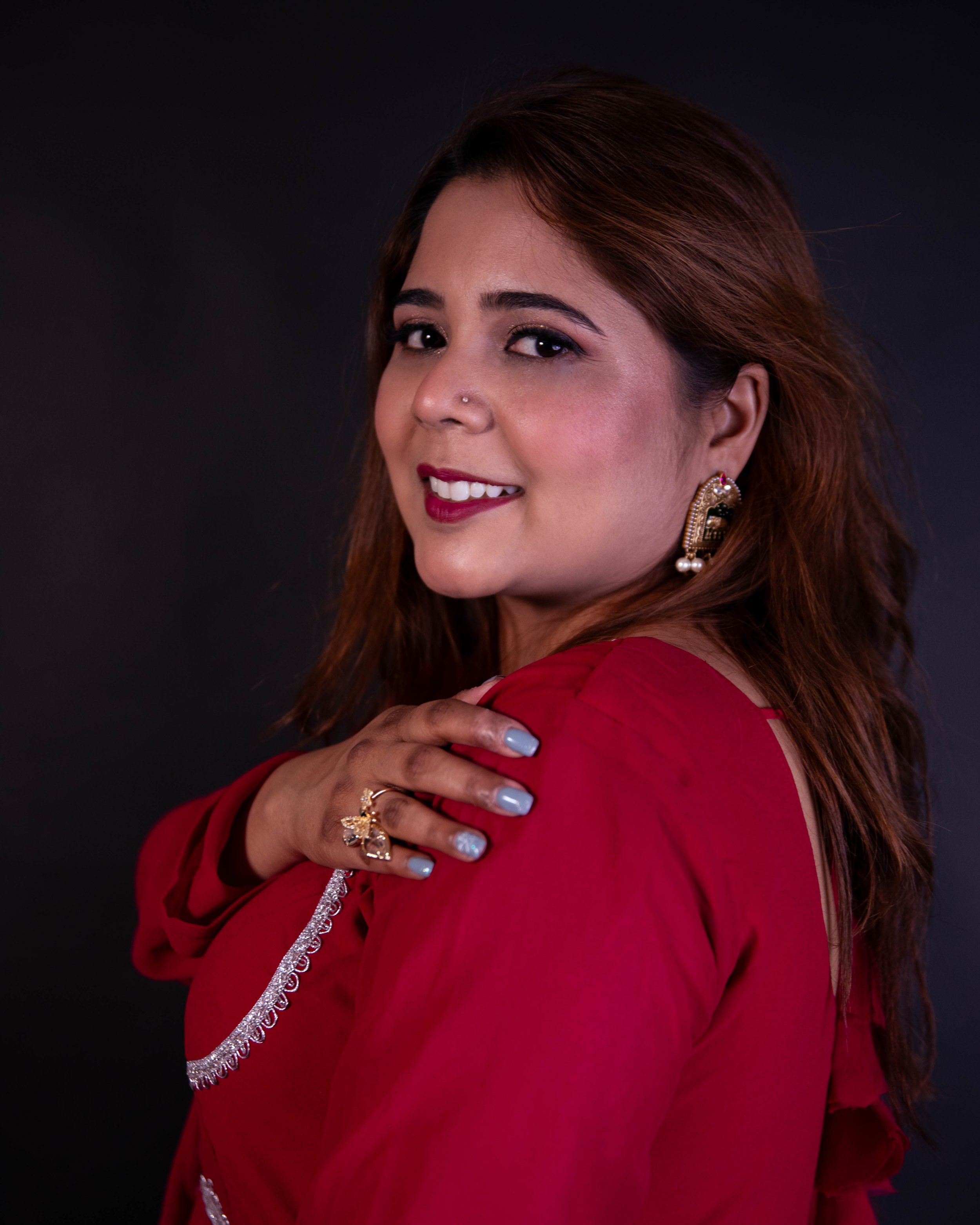 Close-up of a woman with brown hair styled in loose waves, wearing traditional jewelry including earrings with pearls and a ring, and a red saree with silver embroidery, smiling and looking over her shoulder against a dark background.