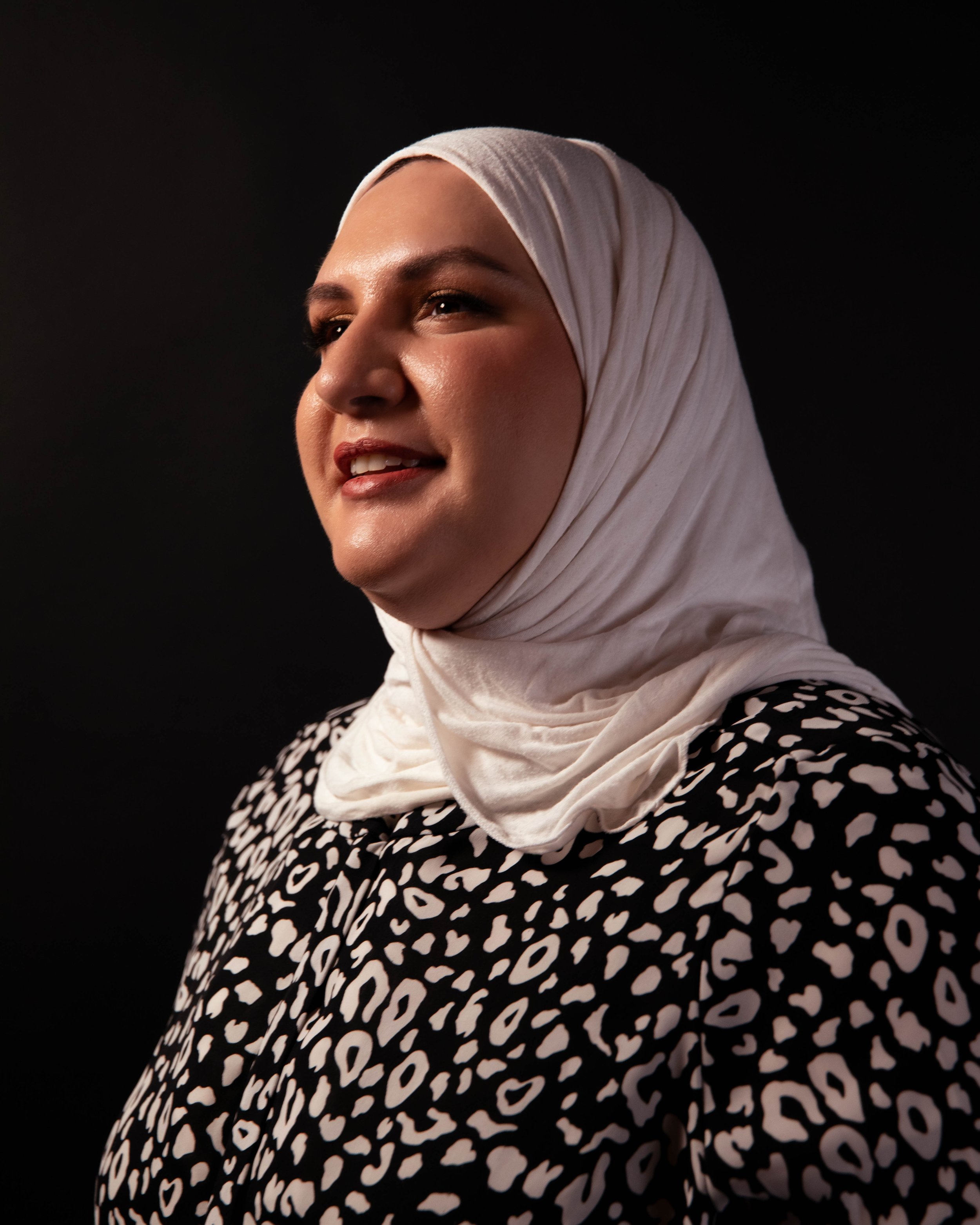A woman wearing a white headscarf, smiling, dressed in a black and white patterned top, against a black background.